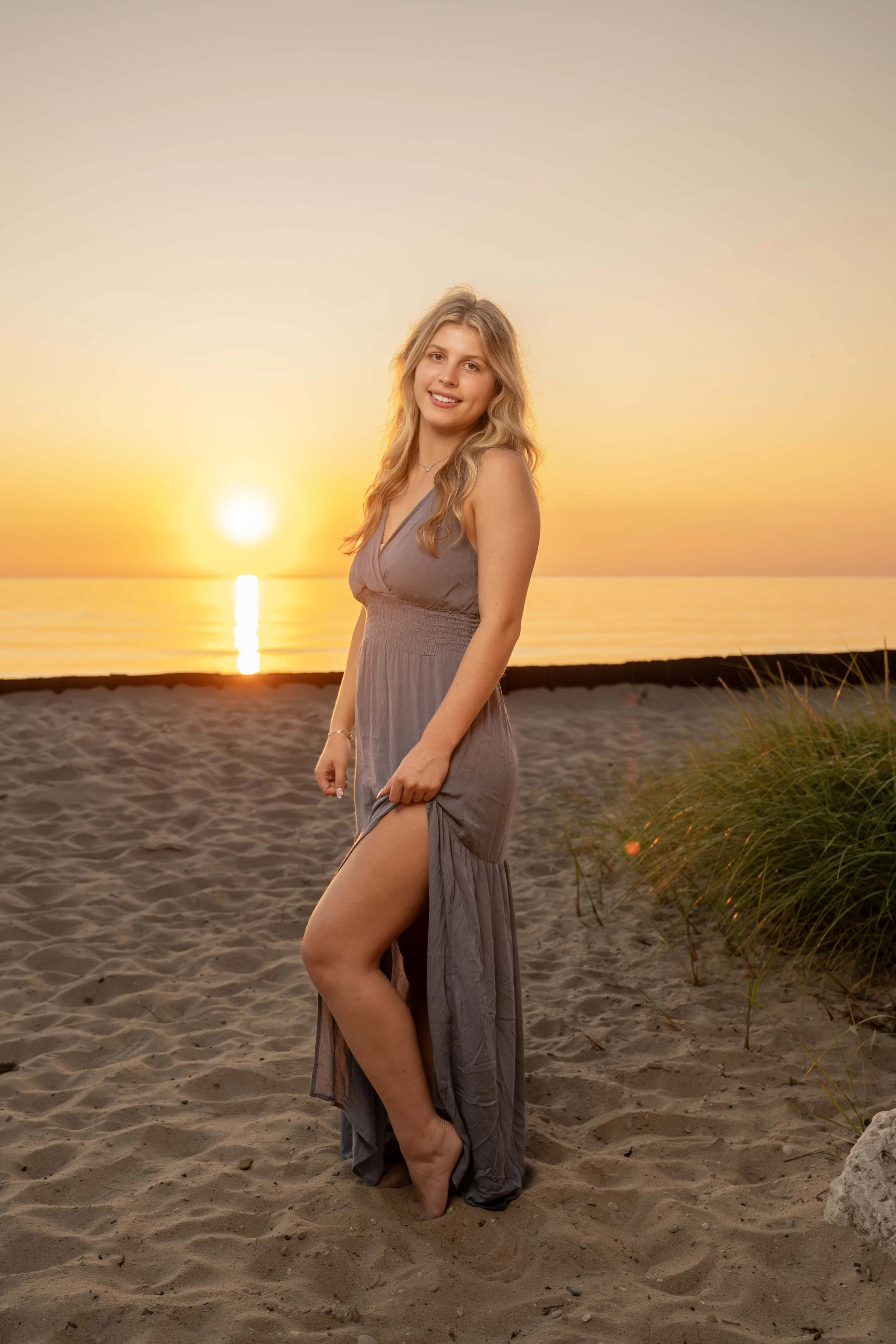 A young woman with long blonde hair standing on a sandy beach at sunset, wearing a gray dress with a slit, smiling at the camera.