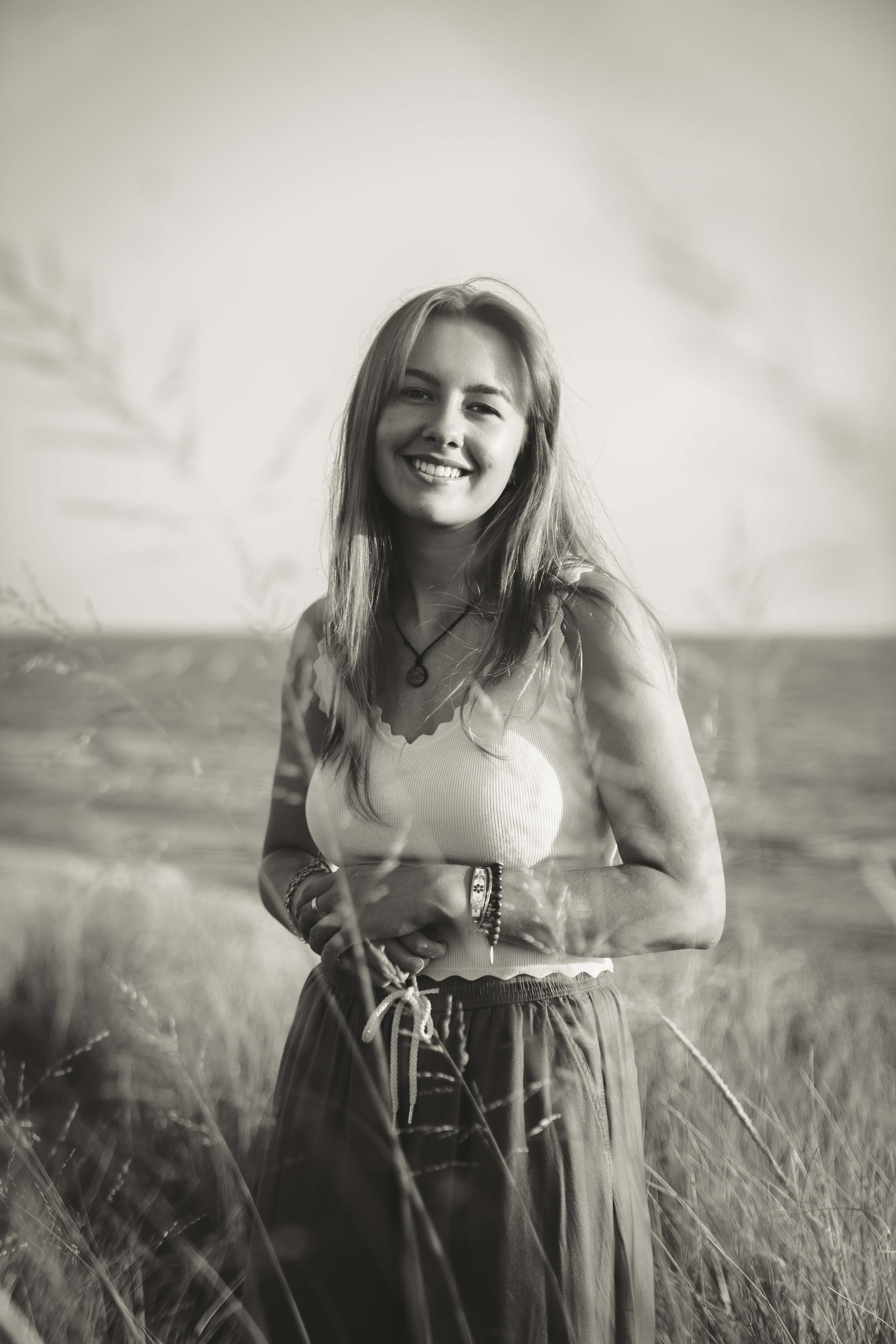 A young woman standing in a grassy field, smiling at the camera, wearing a sleeveless top and loose pants, with long hair, necklaces, and bracelets.