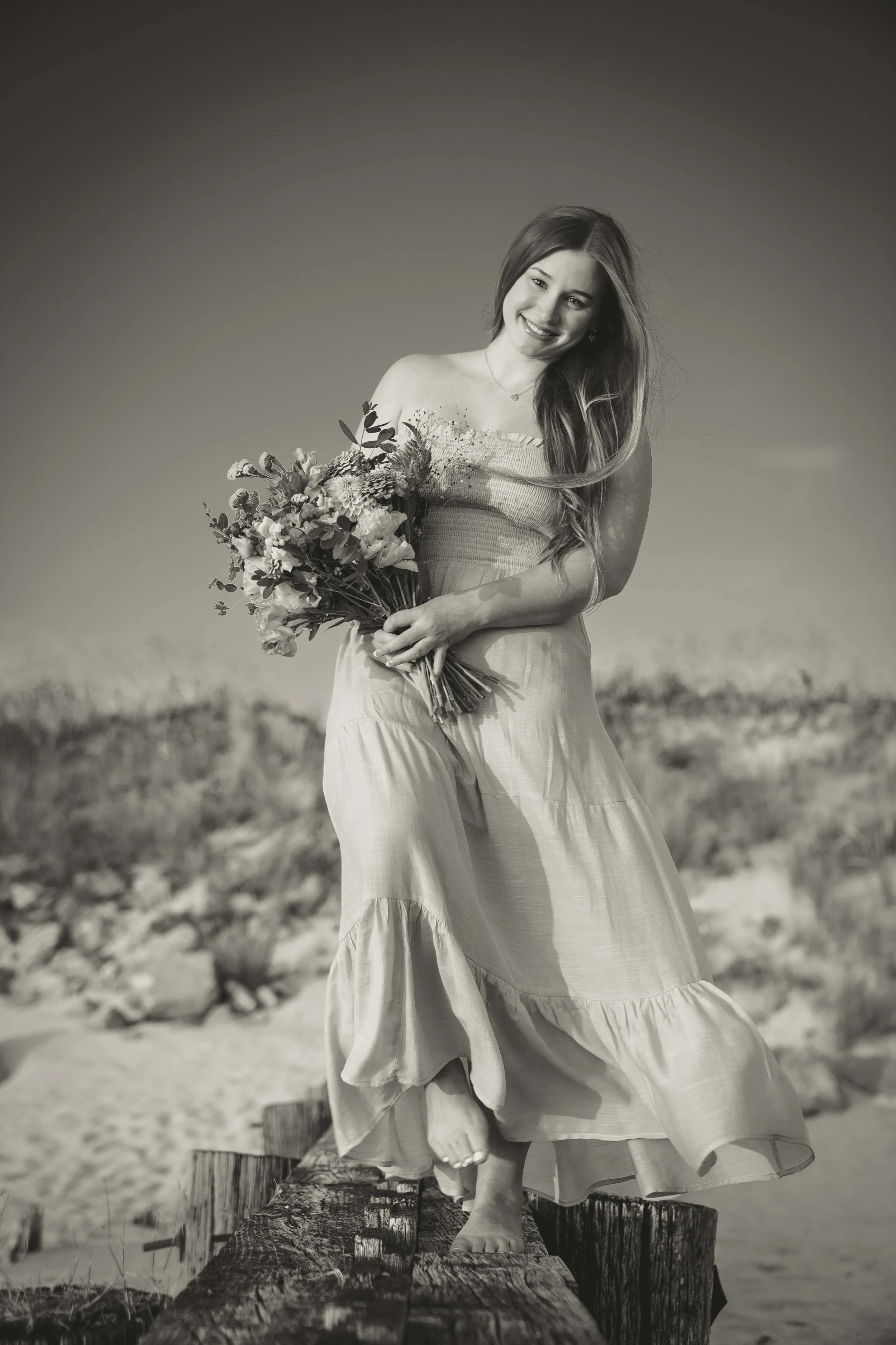 A woman in a long dress holding a bouquet of flowers, standing on a wooden beam outdoors, smiling and looking at the camera, in black and white.