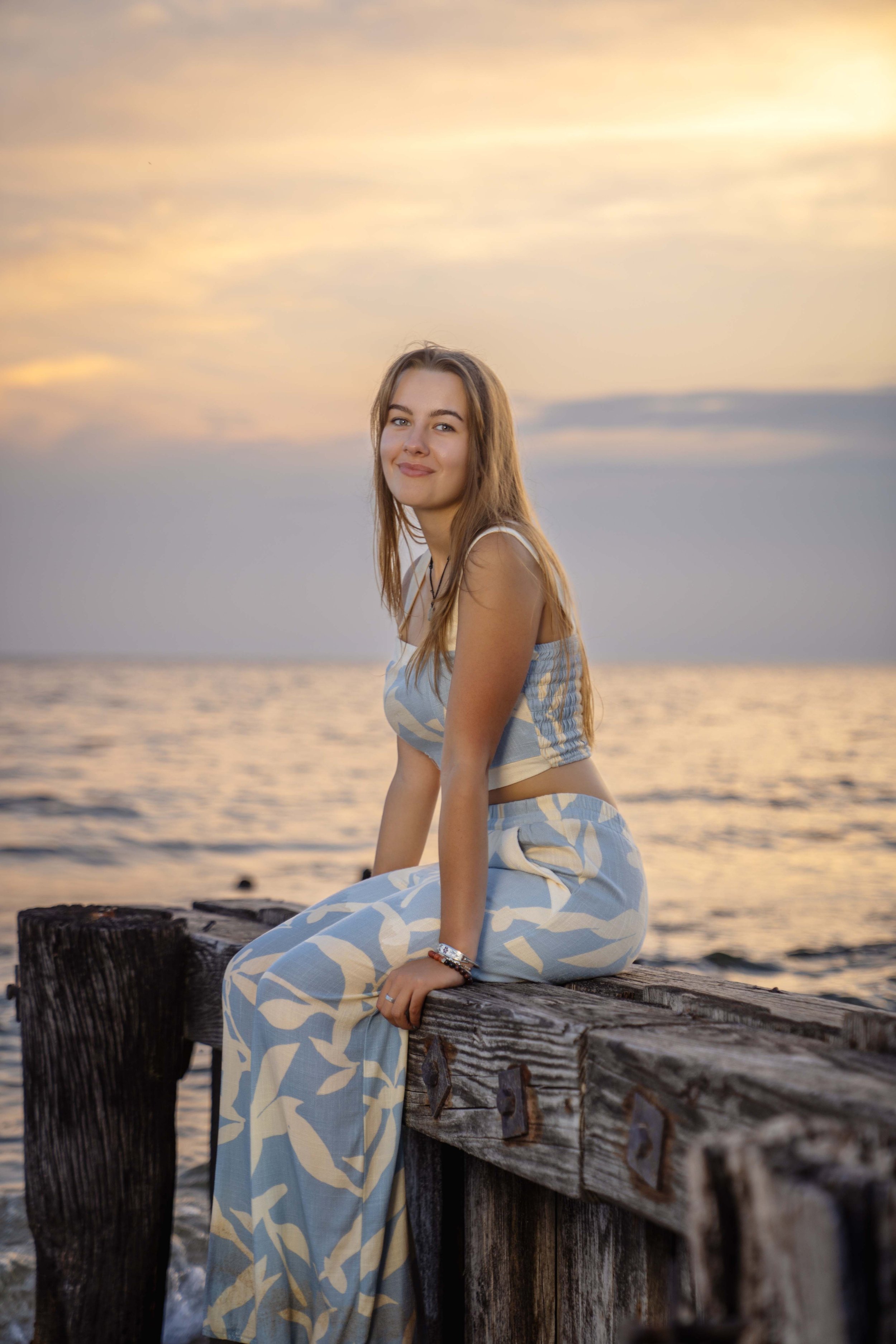 A young woman with long light brown hair sitting on a wooden pier by the ocean during sunset, wearing a matching blue and cream patterned outfit, smiling at the camera.