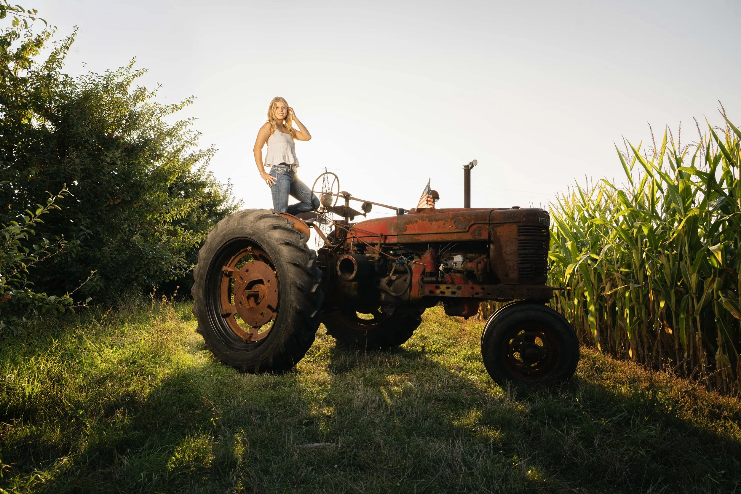 A young woman with long blonde hair wearing a white tank top and jeans, sitting on an old rusty tractor in a rural field, with tall green grass and crops on either side, under clear sky.