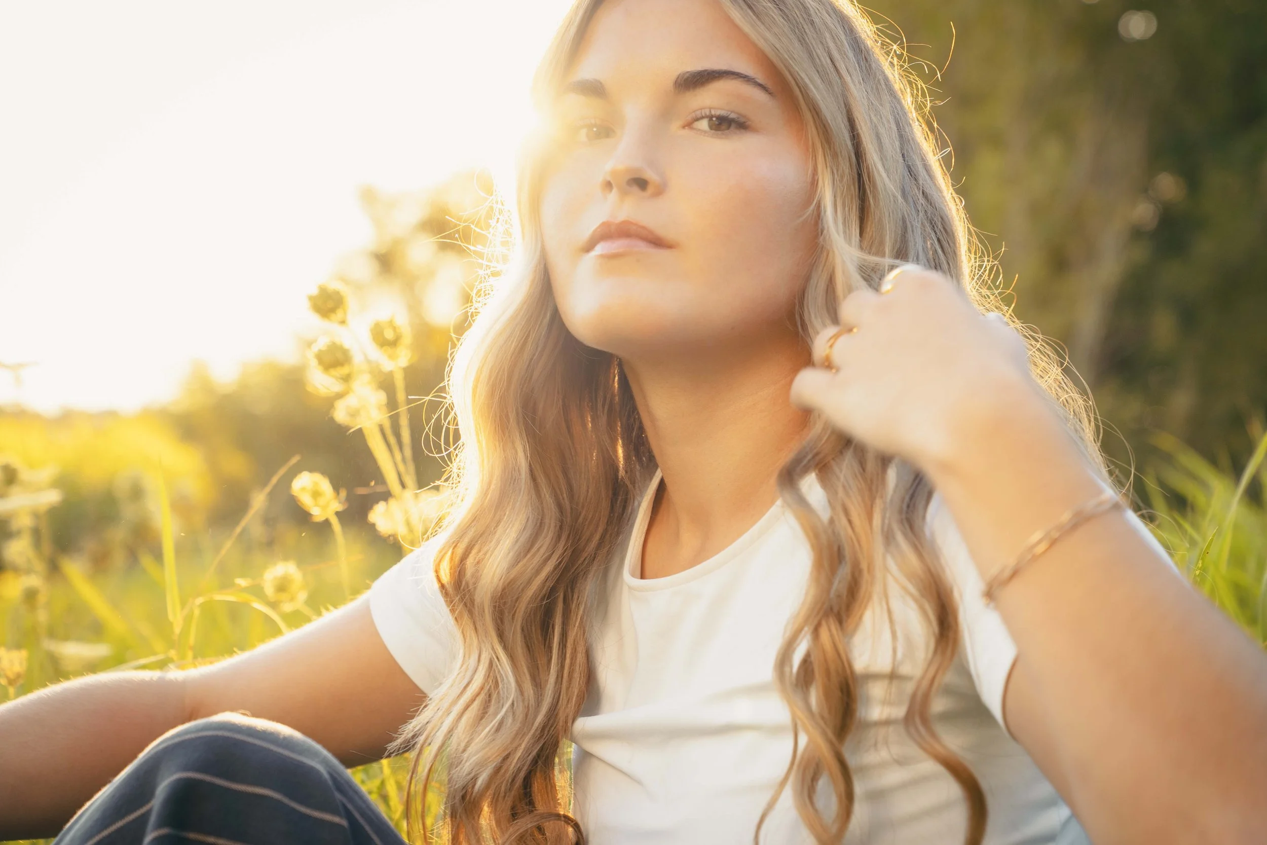 Young woman with long wavy hair sitting in a field at sunset, looking at the camera, wearing a white t-shirt and jewelry, with sunlight behind her.