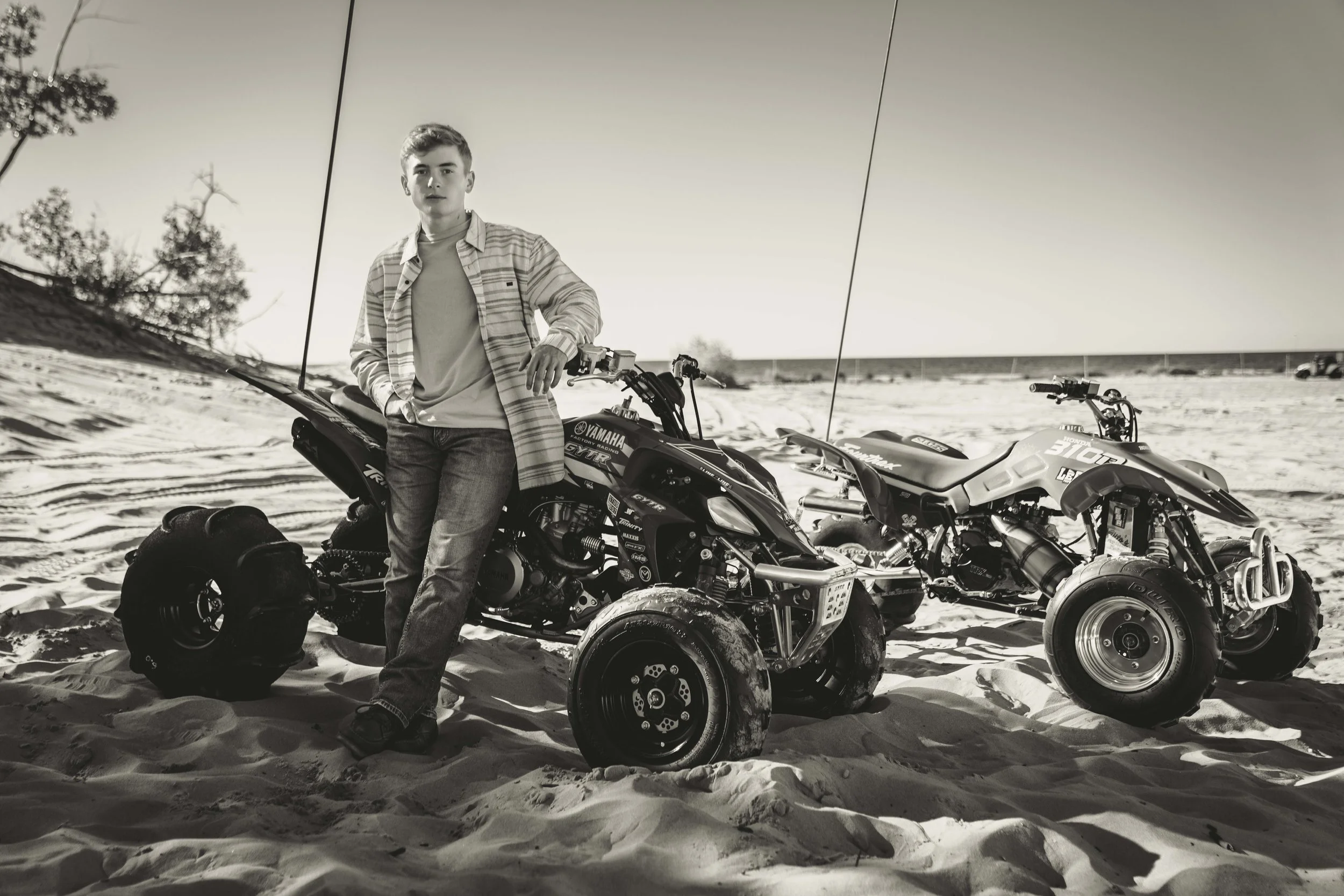 A young man standing next to two all-terrain vehicles (ATVs) on a sandy beach, with trees in the background and a bright sky.