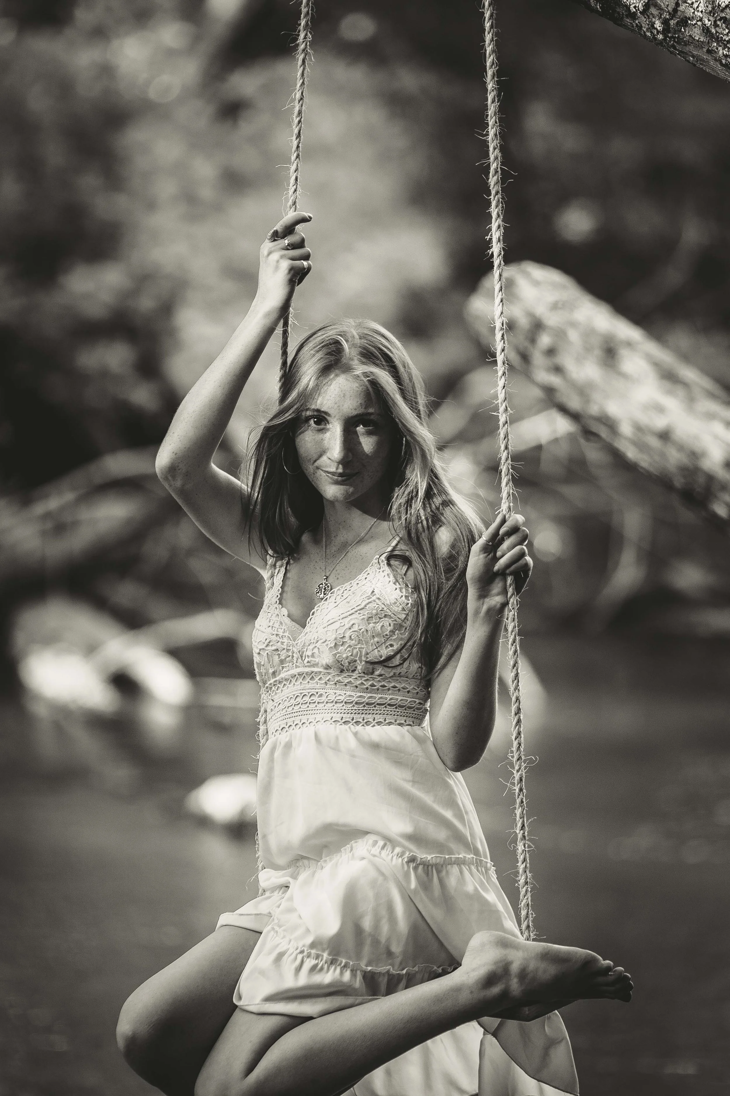 A young woman with long hair hanging on a swing outdoors, smiling slightly, in a black-and-white photo.