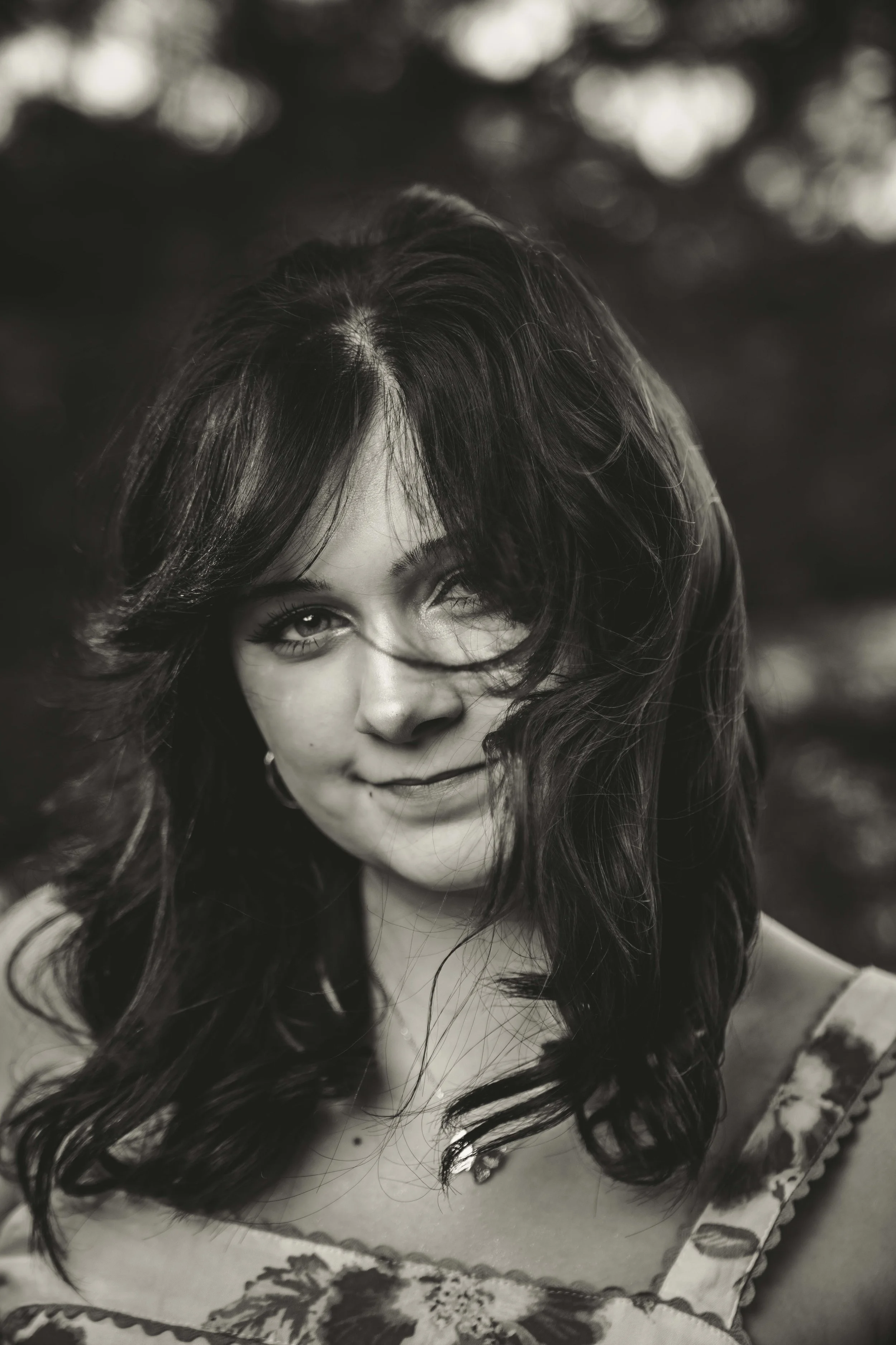 A black and white portrait of a woman with wavy hair partially covering her face, smiling slightly, outdoors with blurred trees in the background.