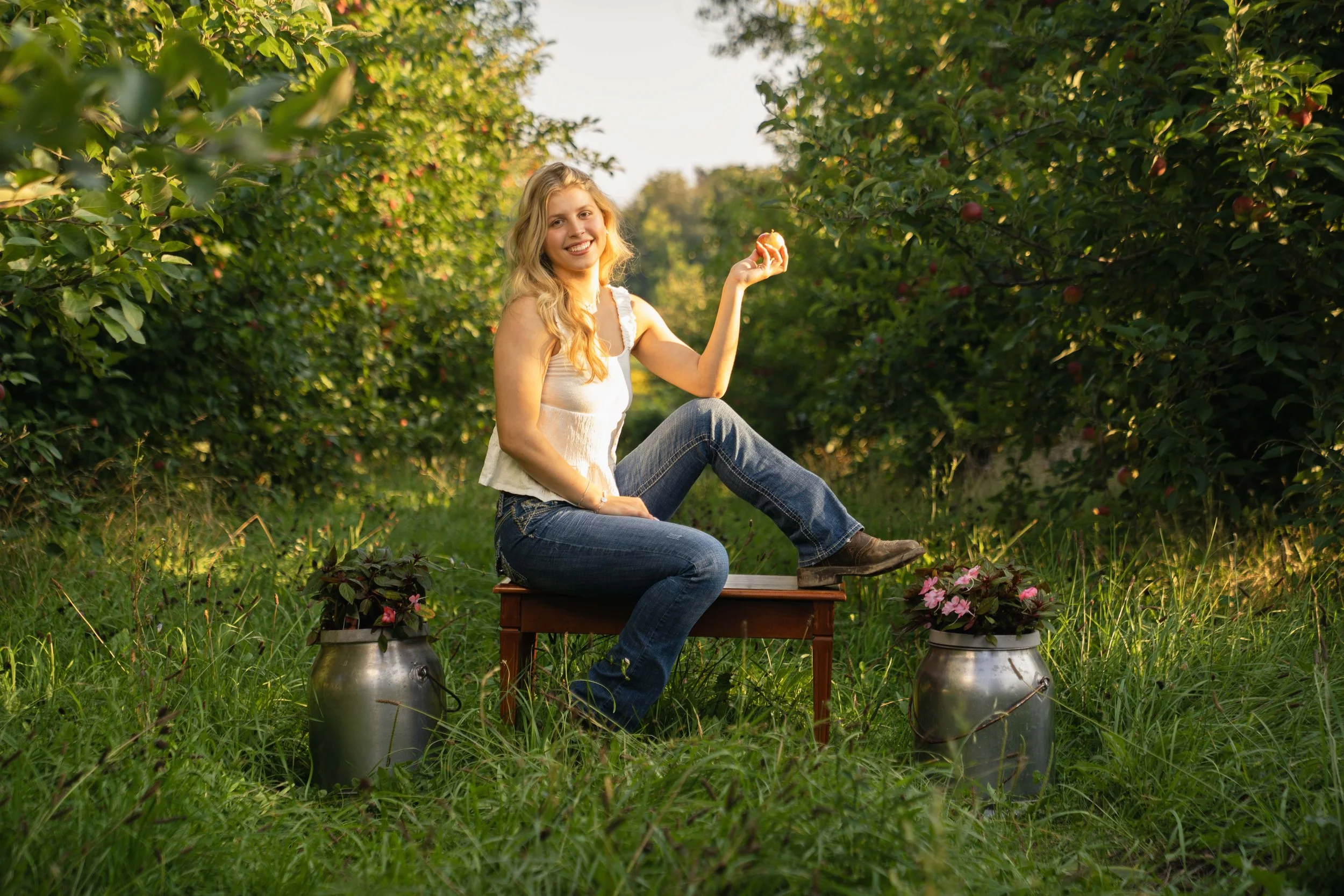 A young woman with long blonde hair, wearing a white sleeveless top and jeans, sitting on a wooden bench outdoors. She is holding an apple in her right hand, smiling, surrounded by green foliage and pink flowers in metallic pots.