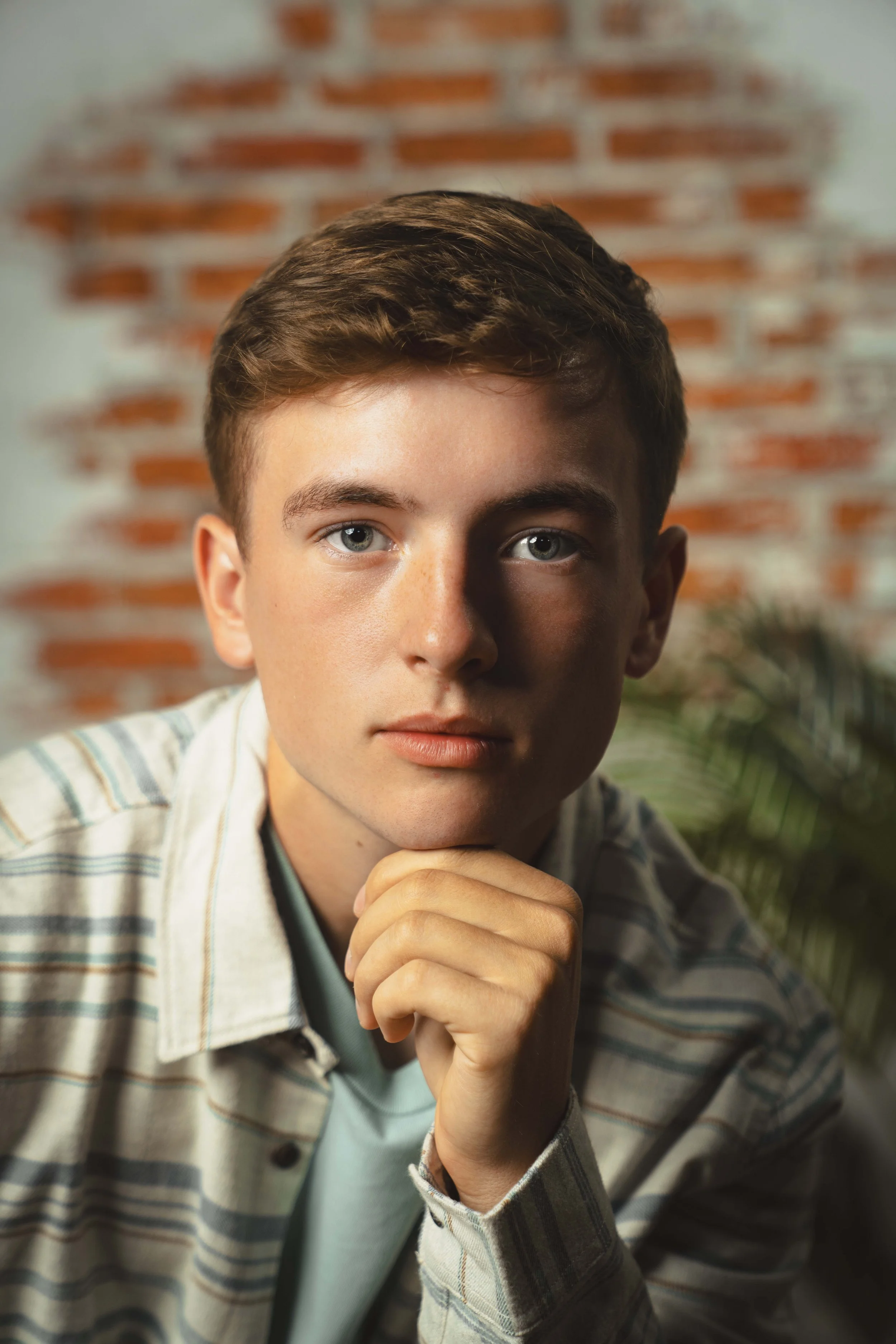 Portrait of a young man with short brown hair, blue eyes, and fair skin, resting his chin on his hand, wearing a plaid shirt over a light blue t-shirt, against a background of a brick wall and a plant.