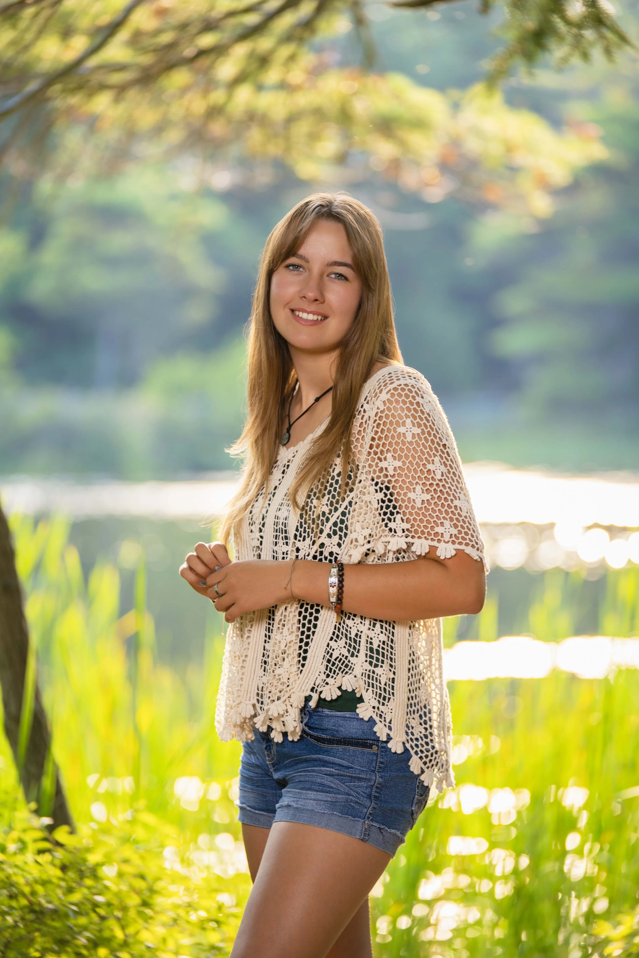 A young woman standing outdoors near a lake, smiling, with trees and greenery in the background, during what appears to be late afternoon or early evening.