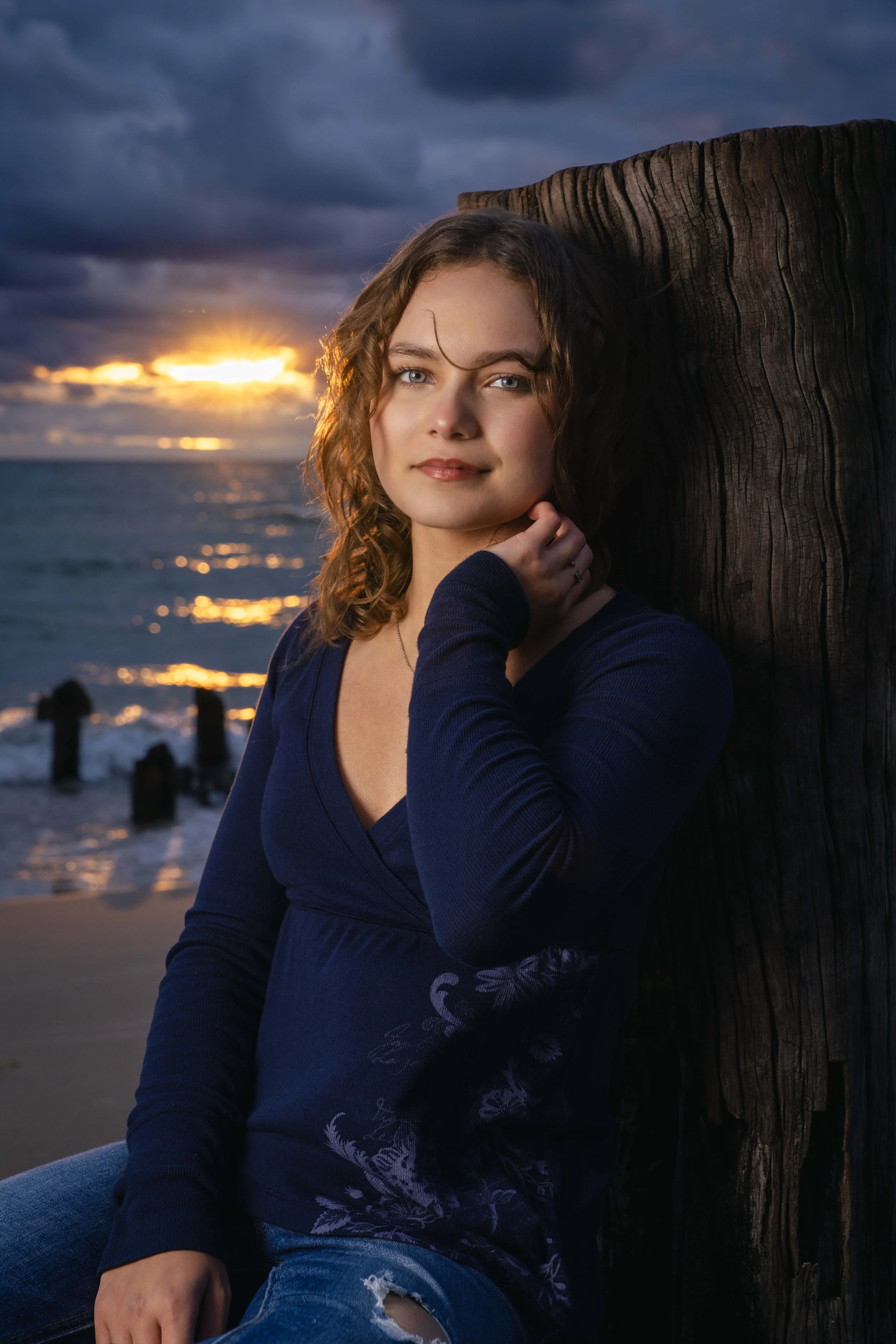 Young woman with wavy hair leaning against a large wooden post at sunset by the ocean.