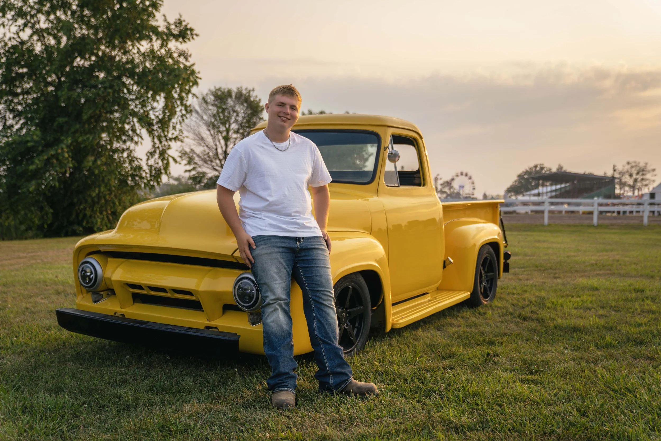 Young man standing with hand on hip in front of a yellow vintage truck on a grassy field at sunset.