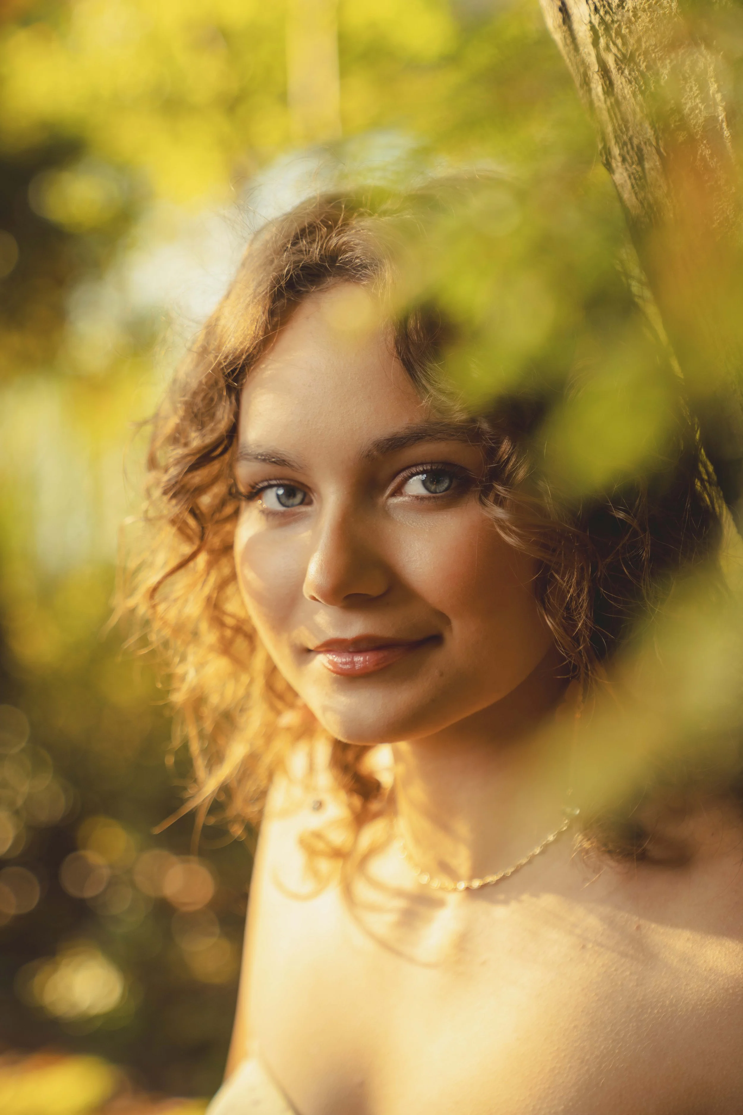 A woman with curly hair and blue eyes, smiling softly, outdoors during golden hour with warm sunlight and blurred foliage in the background.