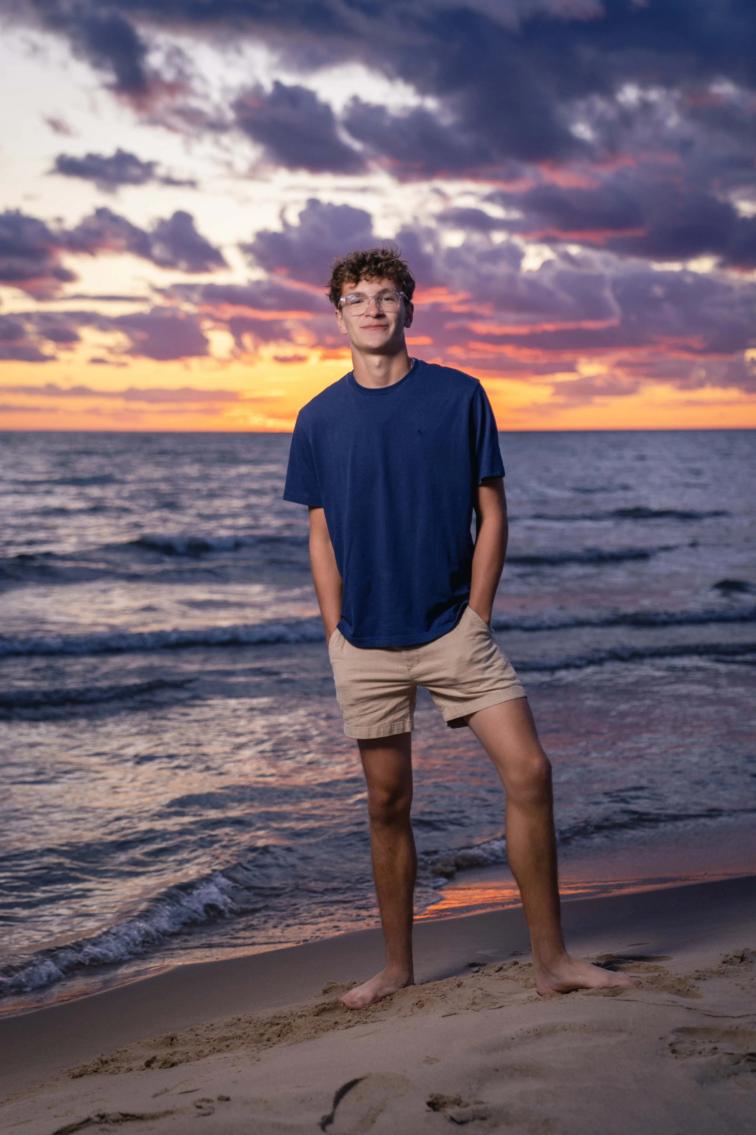 A young man in a blue t-shirt and khaki shorts standing barefoot on the sandy beach at sunset, with clouds and the ocean in the background.