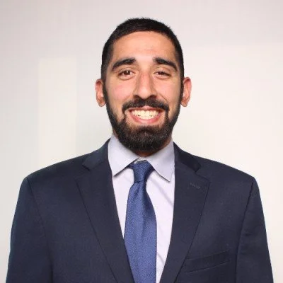 A man with dark hair and a beard wearing a dark suit, white shirt, and blue tie, smiling against a plain light-colored background.