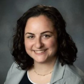 A smiling woman with dark curly hair, wearing a light gray blazer over a dark top, against a dark background.