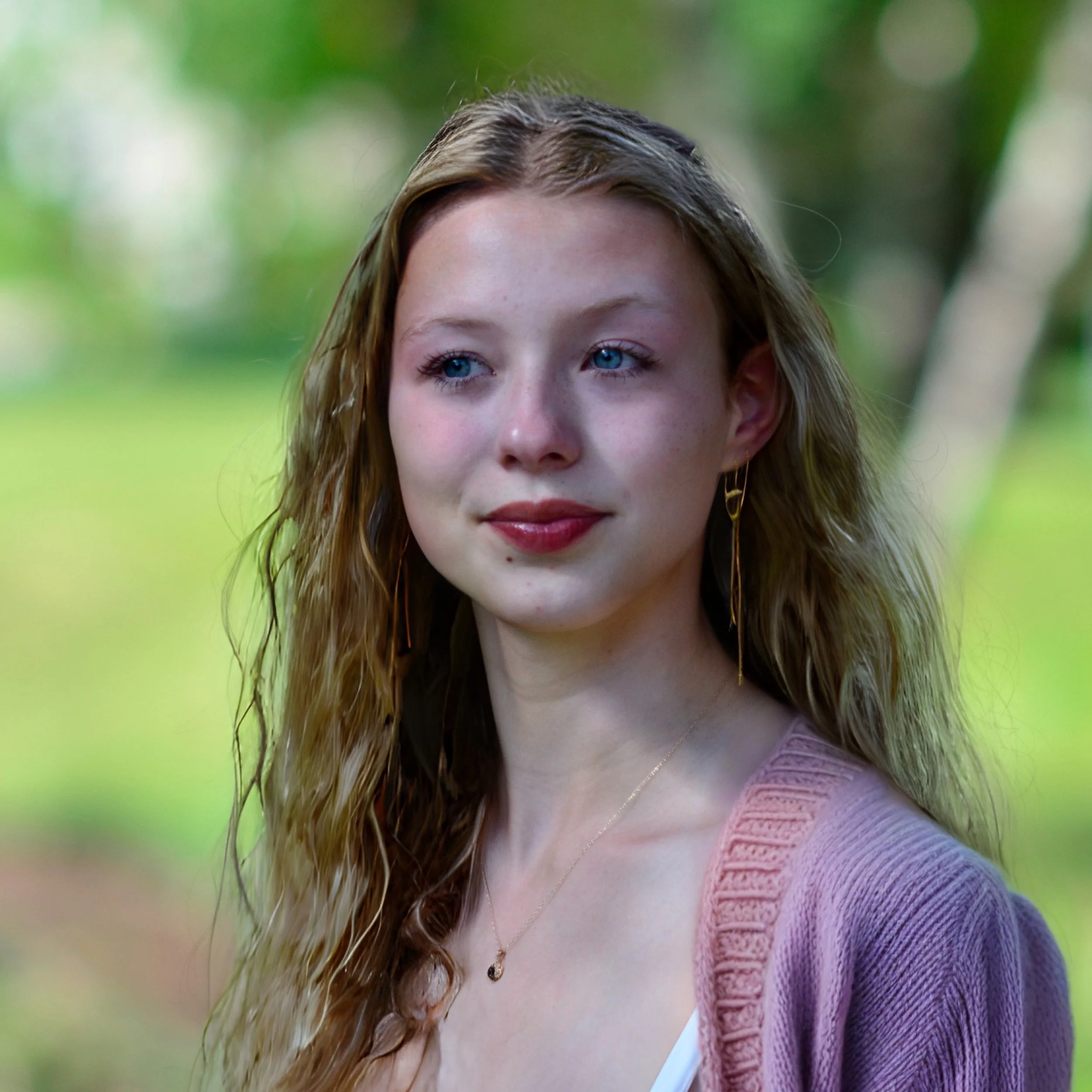 A young woman with long, wavy blonde hair and blue eyes, wearing gold earrings and a pink cardigan, standing outdoors with a blurred green background.