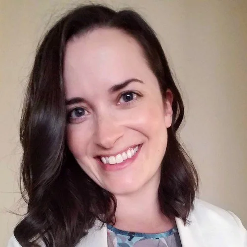 A woman with shoulder-length dark brown hair, smiling, wearing a white blazer and a patterned top, standing against a beige wall.