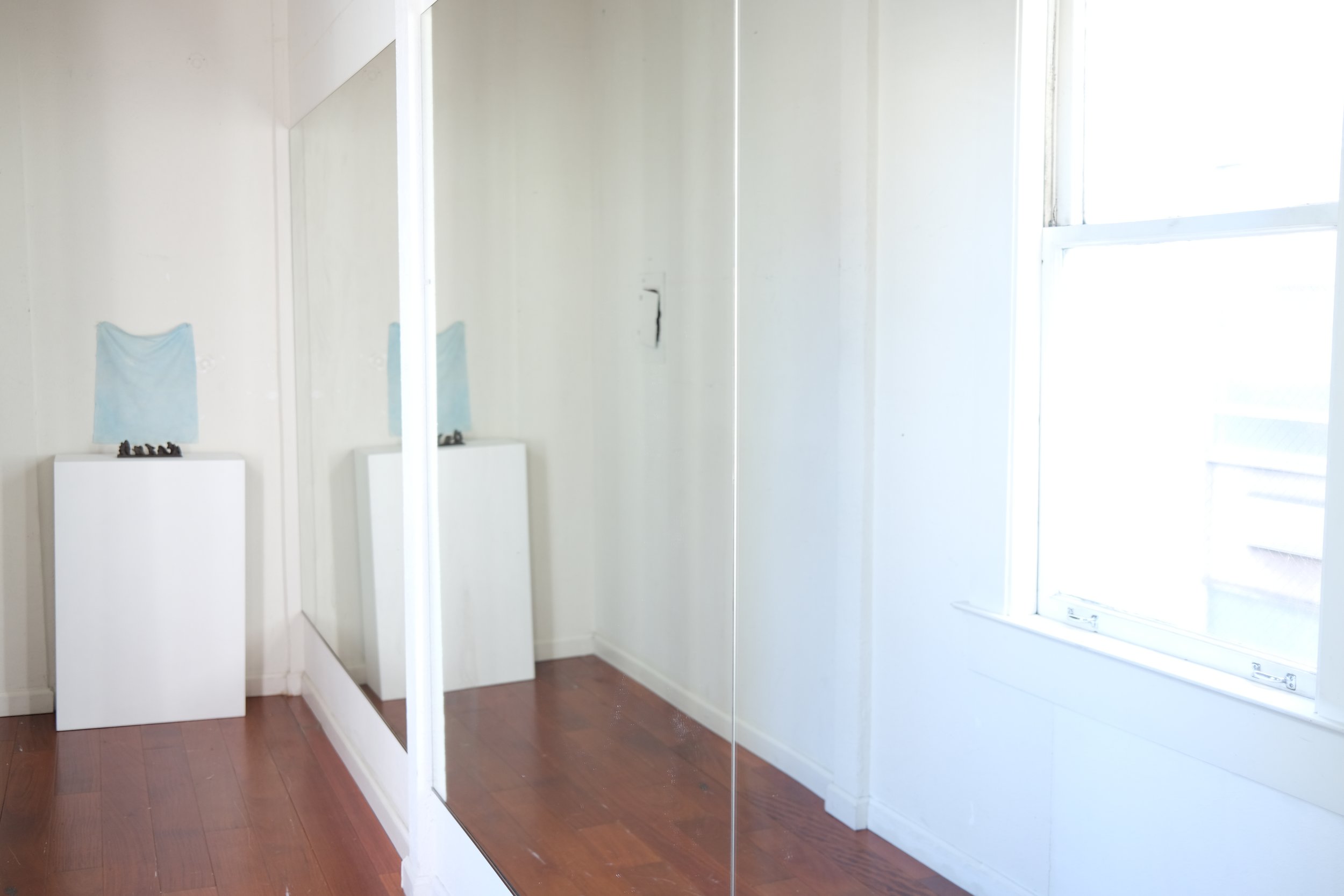 A corner of a room with a wooden floor, a mirror reflecting part of the room, a window letting in light, and a pedestal with a blue cloth draped on it, decorated with small objects.