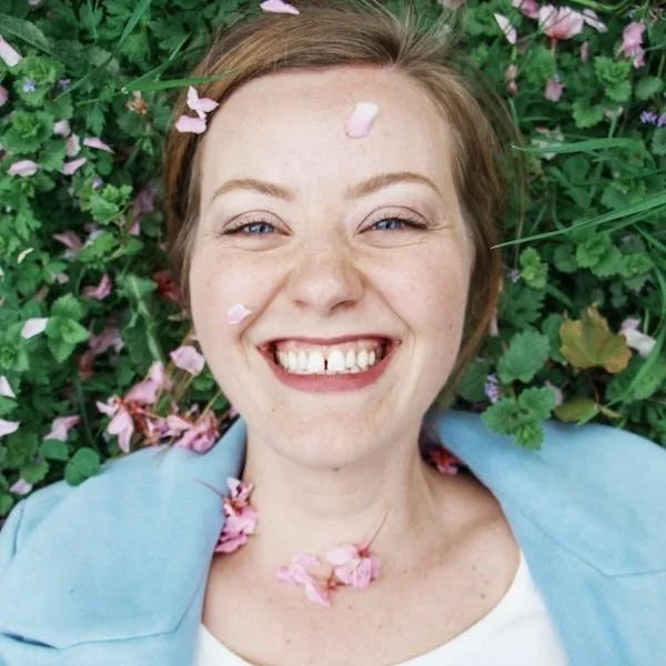 A smiling woman with light skin and light brown hair lying on grass with pink flower petals and green leaves, wearing a light blue jacket and white shirt.