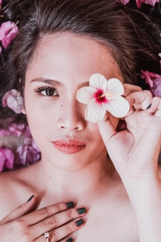 A woman lying down with flowers around her, holding a large white flower with a pink center over one eye.