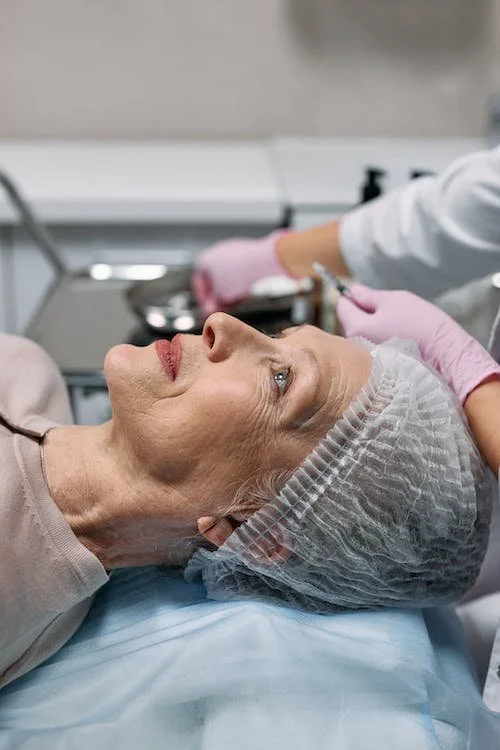 A woman lying on a medical bed with a surgical cap on her head, receiving an injection from a healthcare professional wearing pink gloves in a clinical setting.