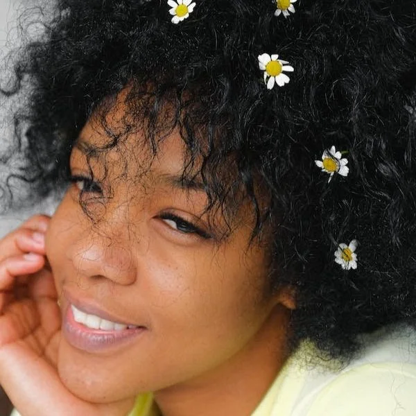 Close-up portrait of a smiling woman with curly black hair decorated with small daisies, resting her head on her hand.