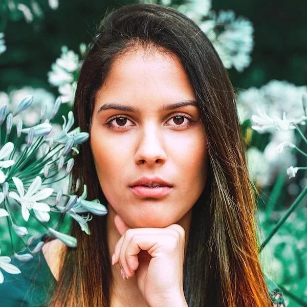 A young woman with long brown hair, natural makeup, and a neutral expression in front of blooming white flowers and green foliage, resting her chin on her hand.