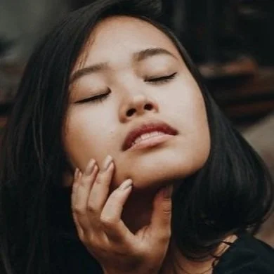 Close-up of a woman with dark hair and closed eyes, gently touching her face with her hand.