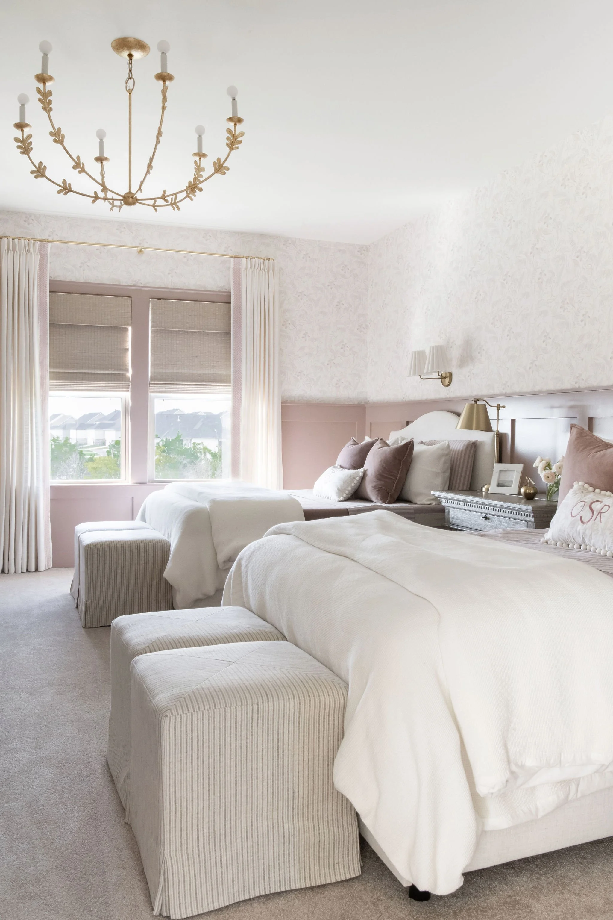 A cozy bedroom with two beds covered in white bedding, facing large windows with cream curtains, and a beige chandelier hanging from the ceiling.