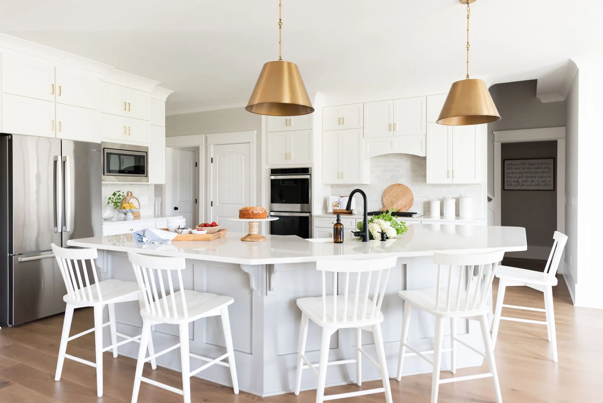 A bright, modern white kitchen with a large island, six white chairs, and gold pendant lights hanging above. The kitchen has stainless steel appliances, including a refrigerator and built-in microwave. Decor includes a cake, flowers, and kitchen esse