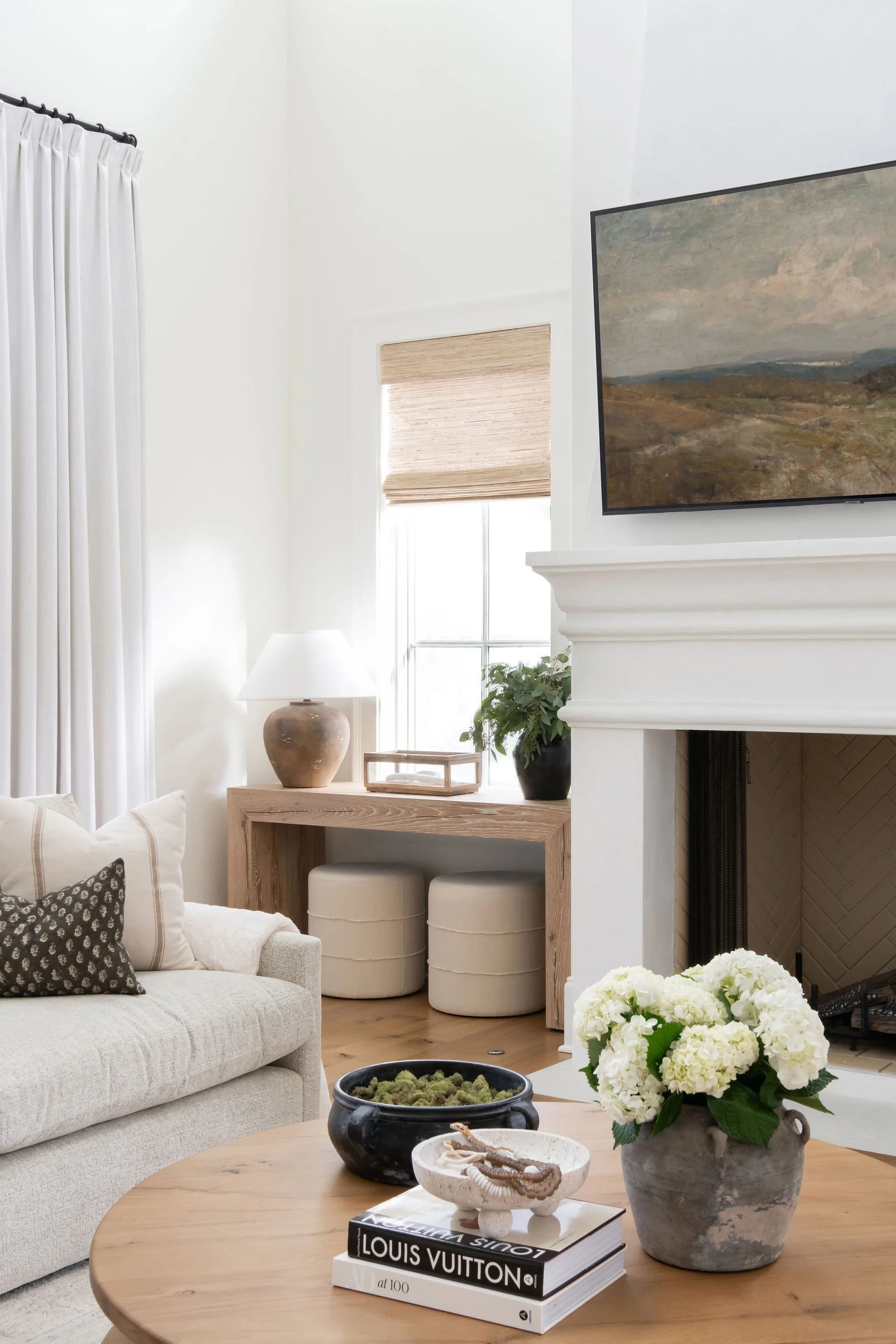Living room with a beige sofa, black and white pillows, wooden coffee table with decorative items and books, white walls, window with woven blinds, fireplace with white mantel, wall-mounted TV, and a flower pot with white flowers.