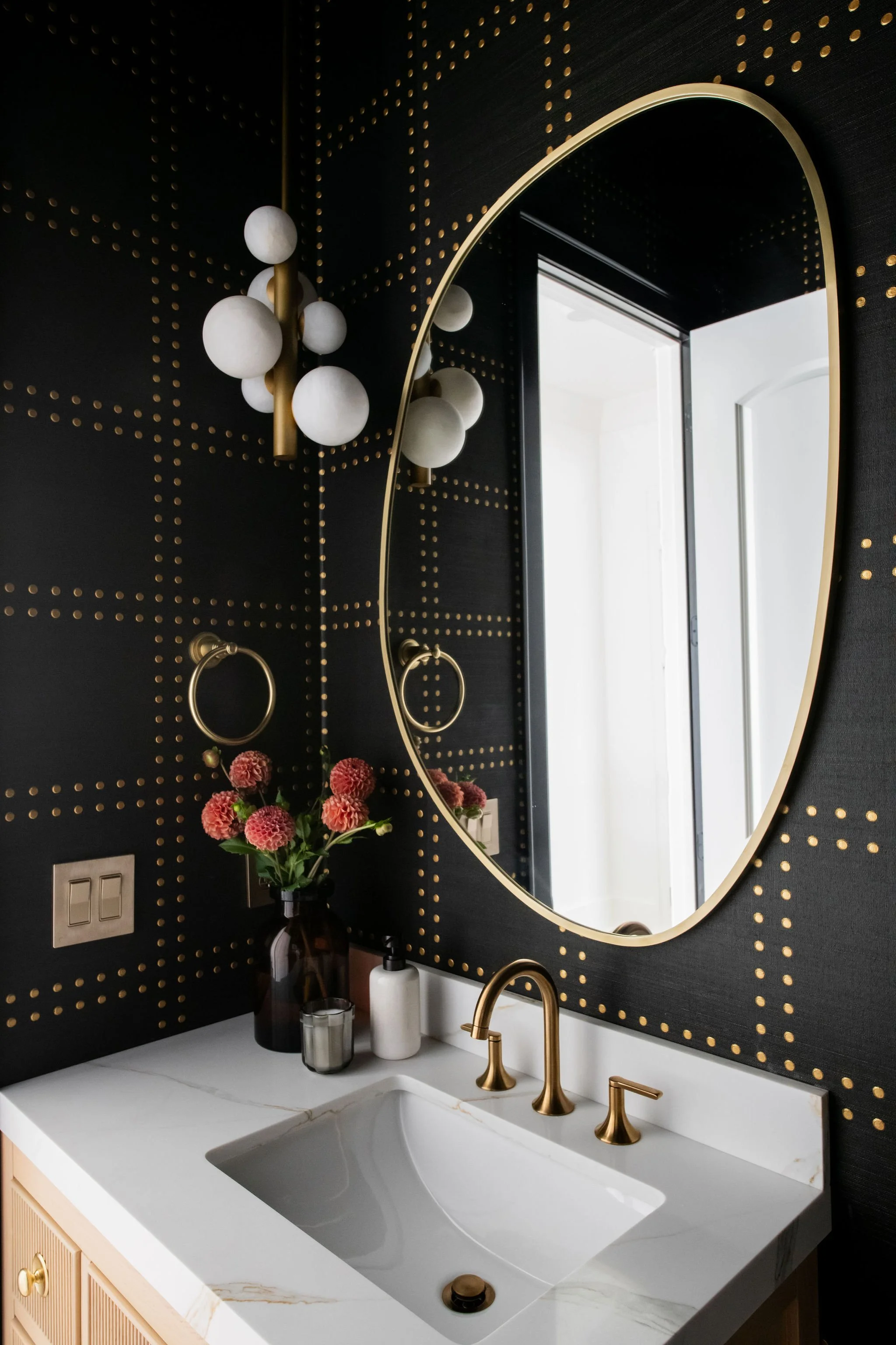 Bathroom vanity with a white marble countertop, a gold faucet, a large oval mirror, black wall with gold dotted pattern, decorative pink flowers in a dark bottle, a soap dispenser, a candle holder, and a wall-mounted gold towel ring.