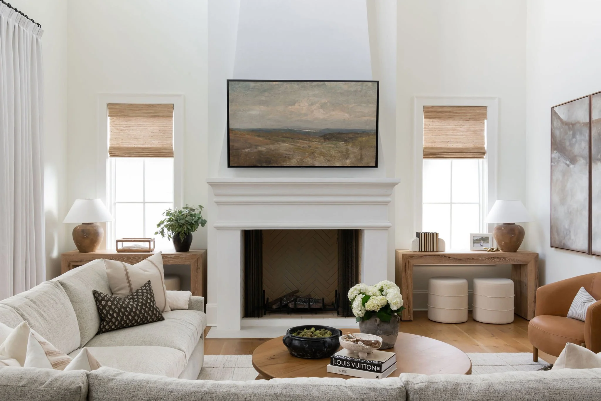 Living room with a white sofa, a round wooden coffee table, a fireplace with a landscape painting above it, and two windows with beige blinds.