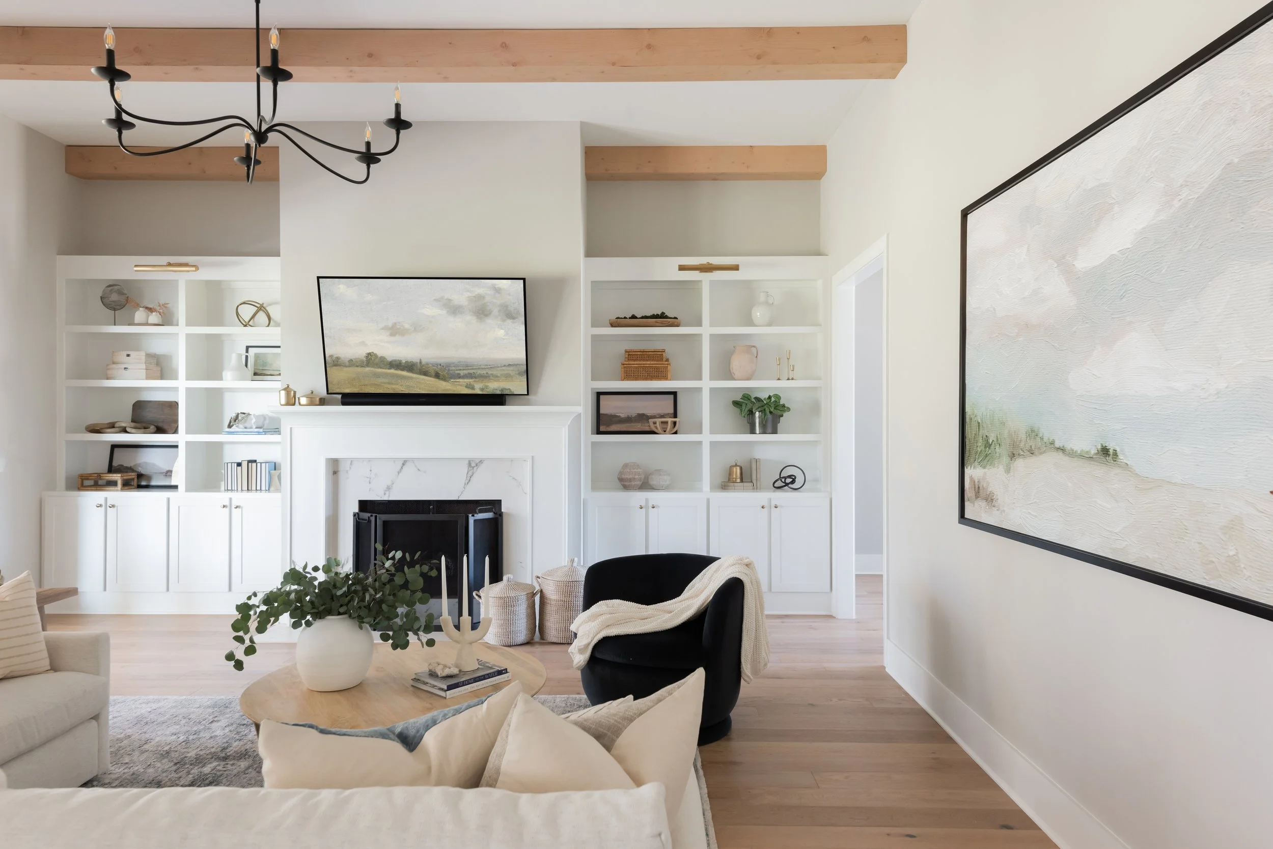 Living room with white built-in shelves, a mounted TV above a fireplace, a black armchair with a blanket, a wooden coffee table with a vase of greenery, and a large abstract painting on the wall