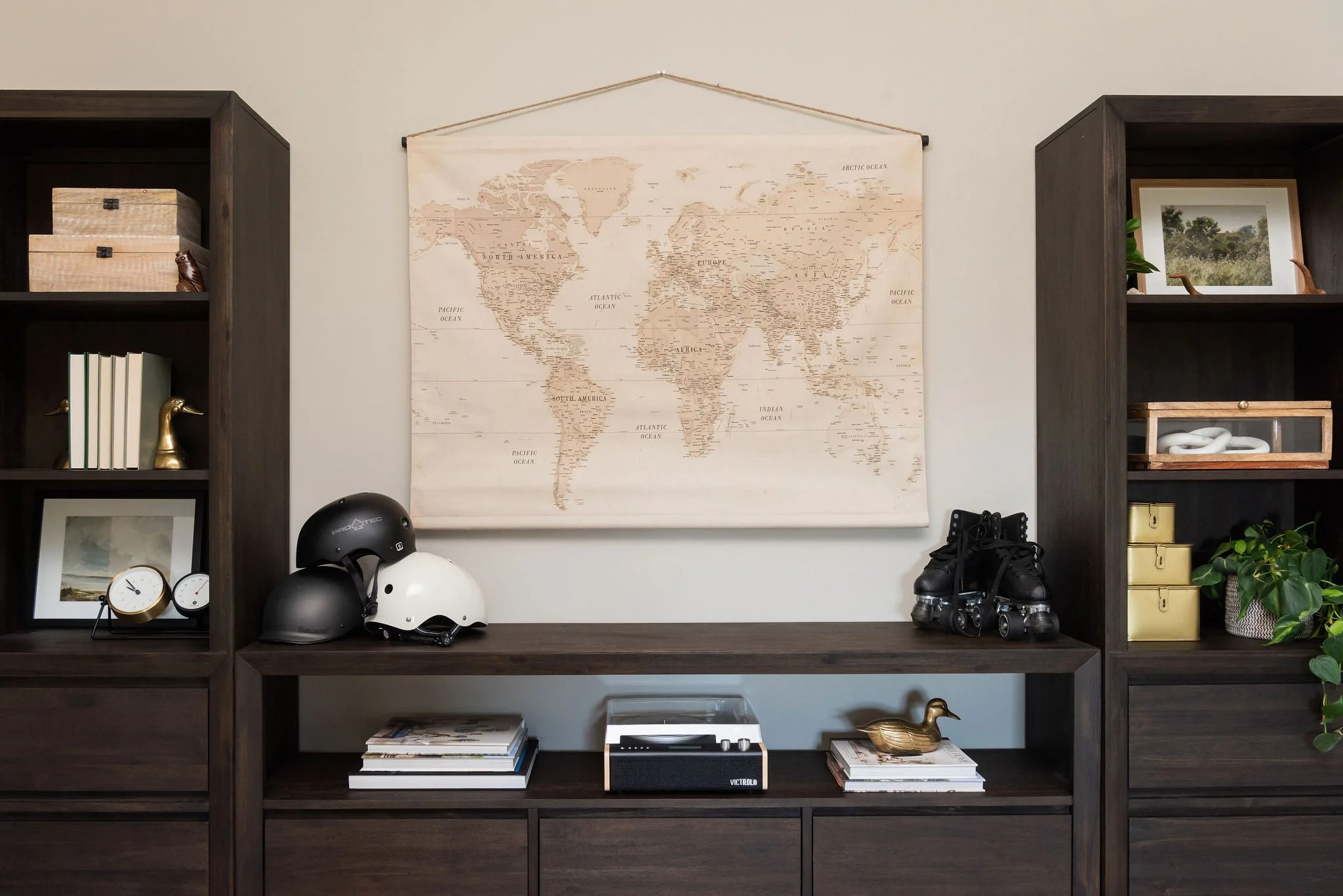 A room with a dark wood storage unit on each side, a beige world map hanging on the wall, three helmets on the shelf, a pair of rollerblades, a small clock, books, plants, and a decorative duck figurine.