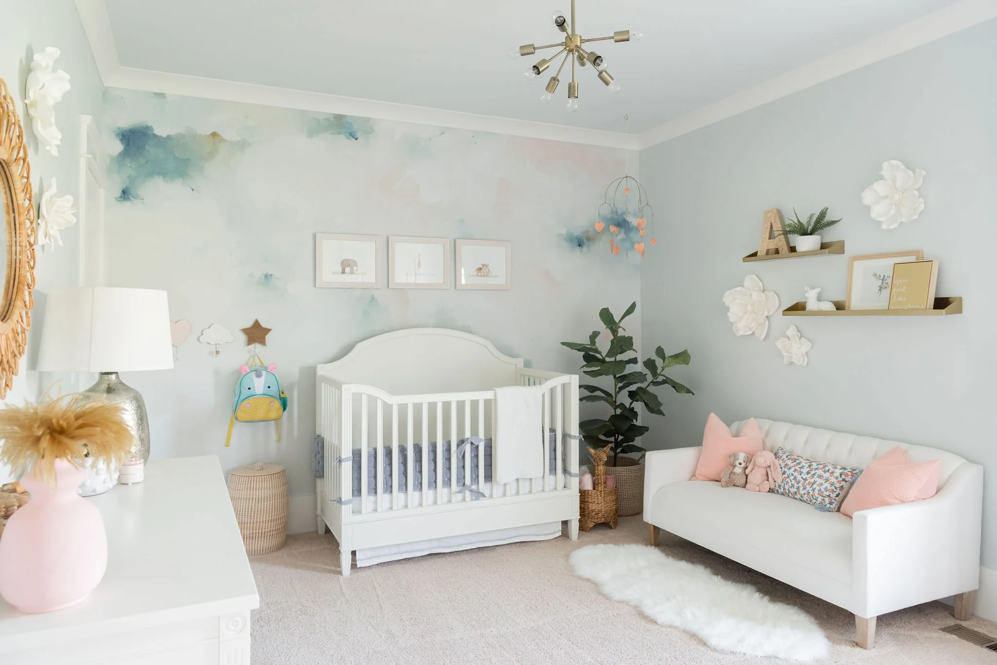 A nursery with a white crib, a white couch with pink pillows, a large green plant, and shelves with decorative items on a light wall with cloud and animal artwork.