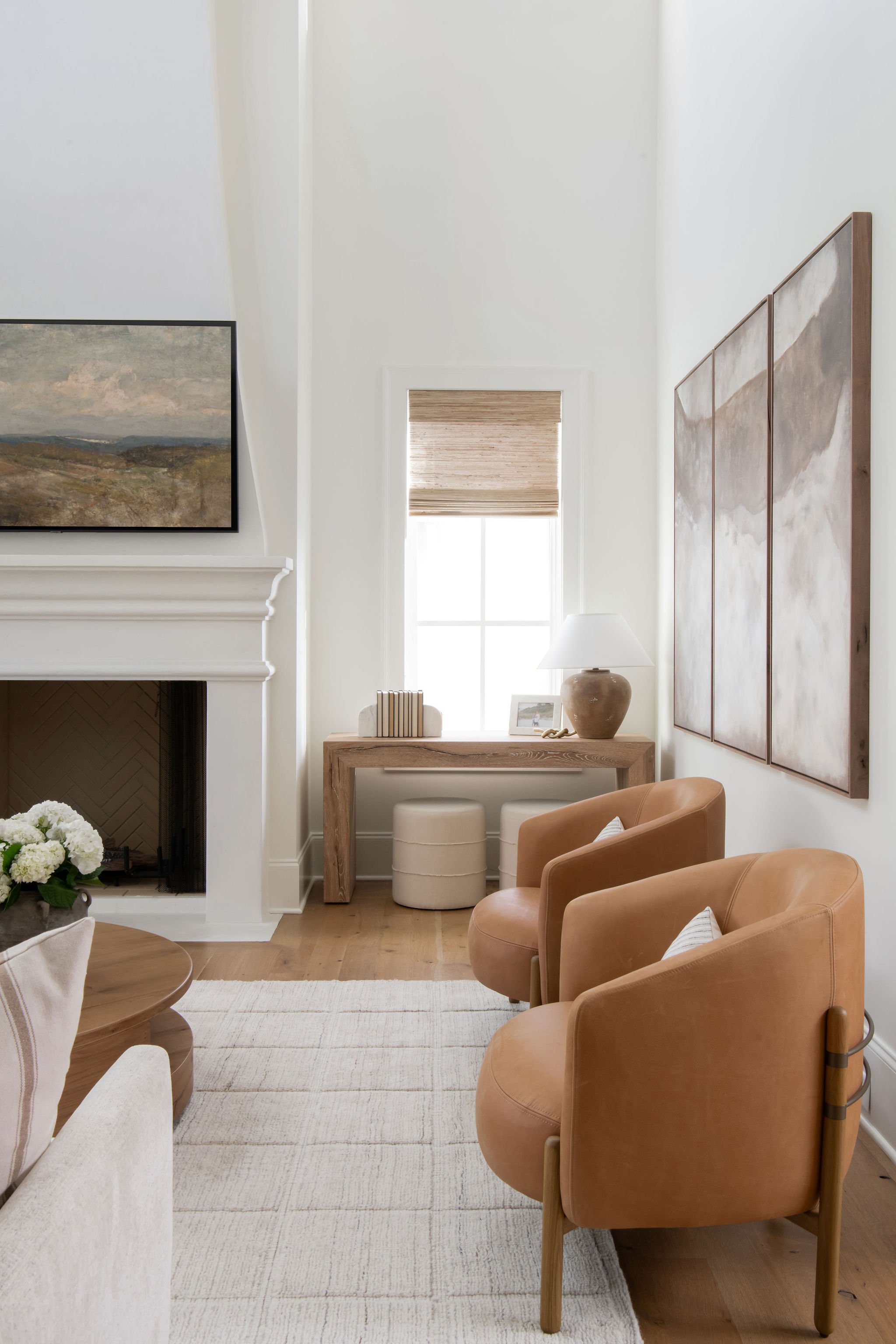 A cozy living room with tan armchairs, a white area rug, a fireplace, a wooden console table with a lamp, books, and framed pictures, a window with a beige shade, and wall art, in a neutral color palette.
