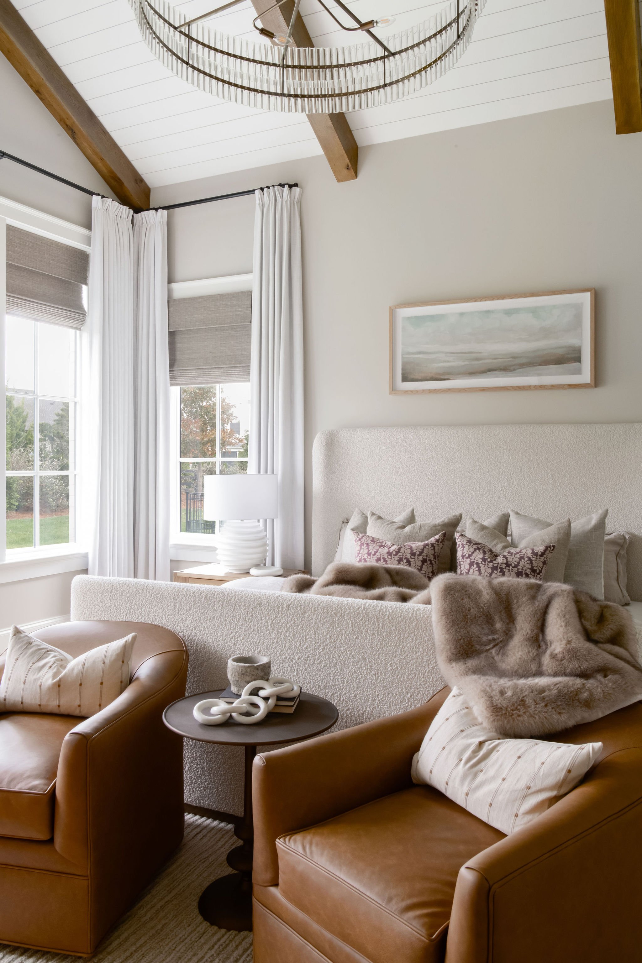 Living room with white walls, large windows with white curtains, a beige upholstered bed or sofa with decorative pillows, a framed landscape painting above it, a side table with a white lamp, two brown leather armchairs with cushions and a fur throw,
