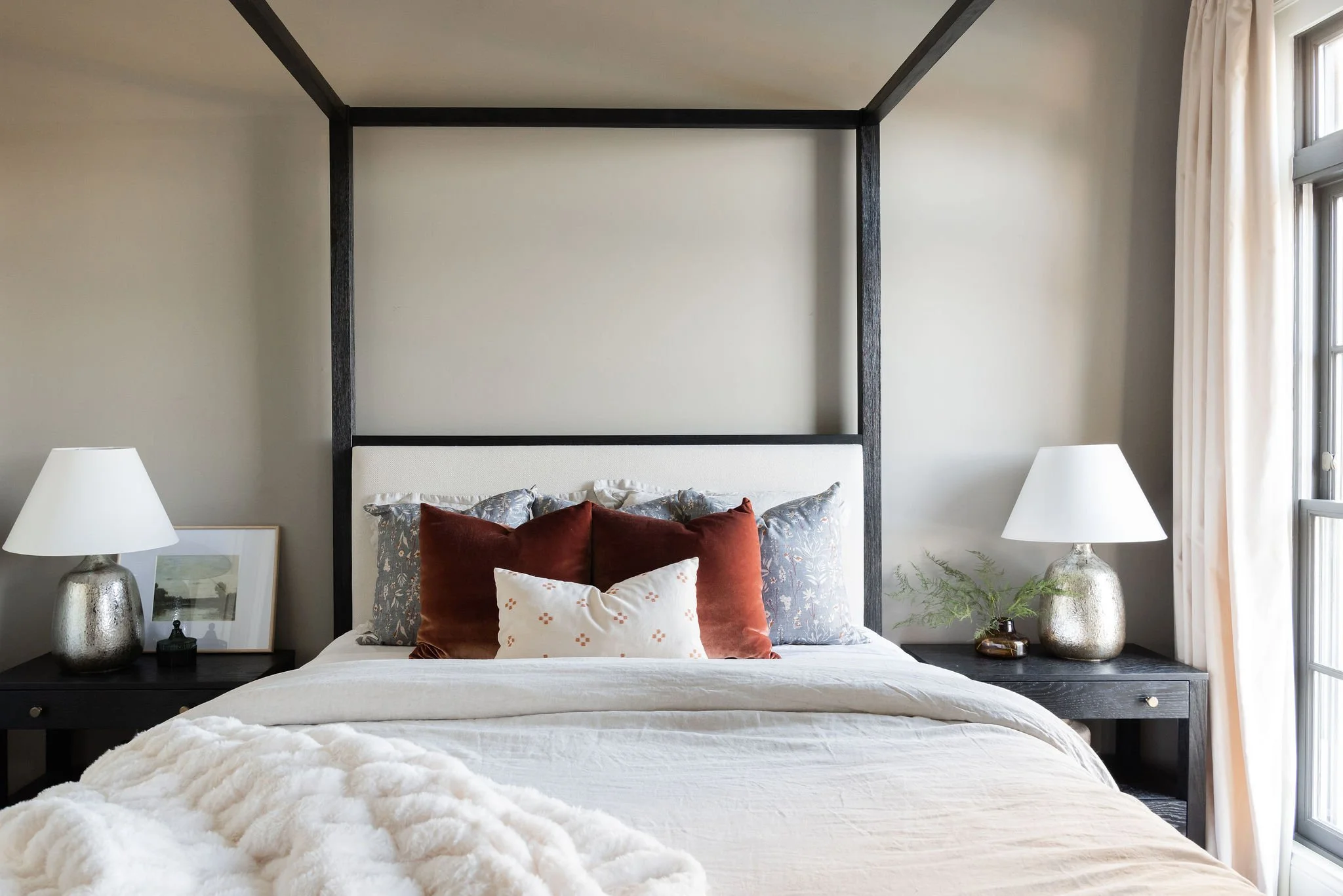 A bedroom with a bed, four pillows, two bedside tables with lamps, and a window with curtains.