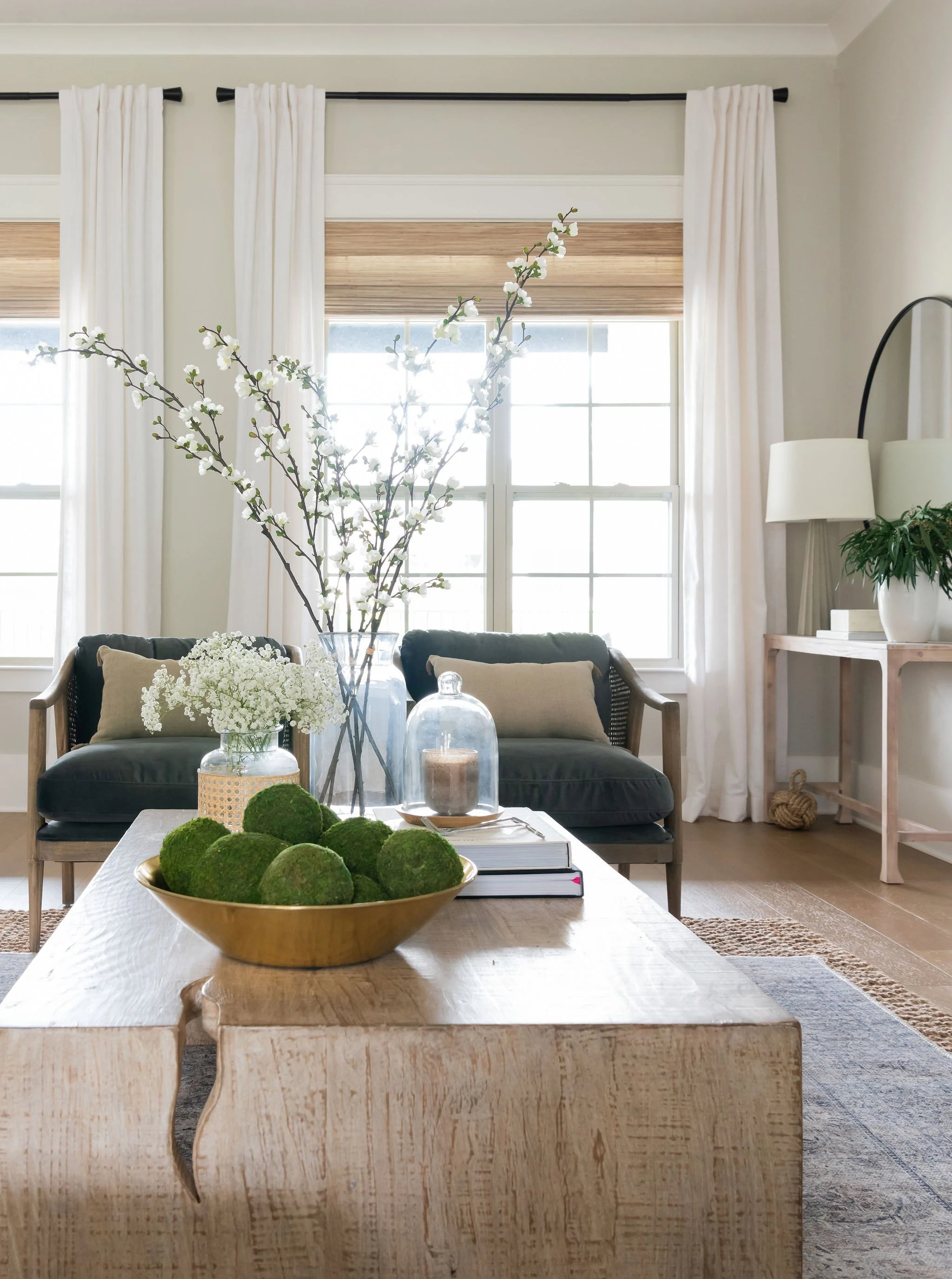 Living room with natural light, decorated with a table of flowers and moss balls, two black cushioned chairs with beige pillows, white curtains, and a white doorway with a mirror and greenery in the background.