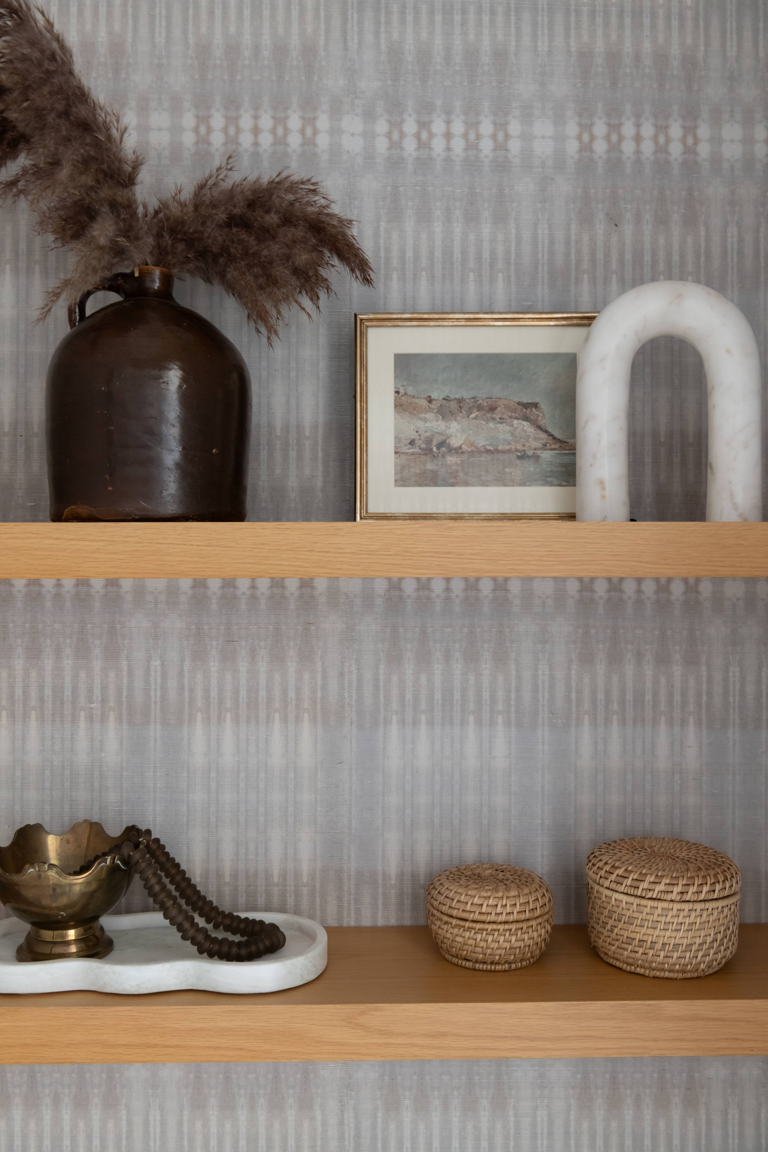 Decorative interior shelving with a dark brown vase with pampas grass, a framed landscape picture, a white marble arch sculpture, brass bowl, bead necklace, and two woven baskets.