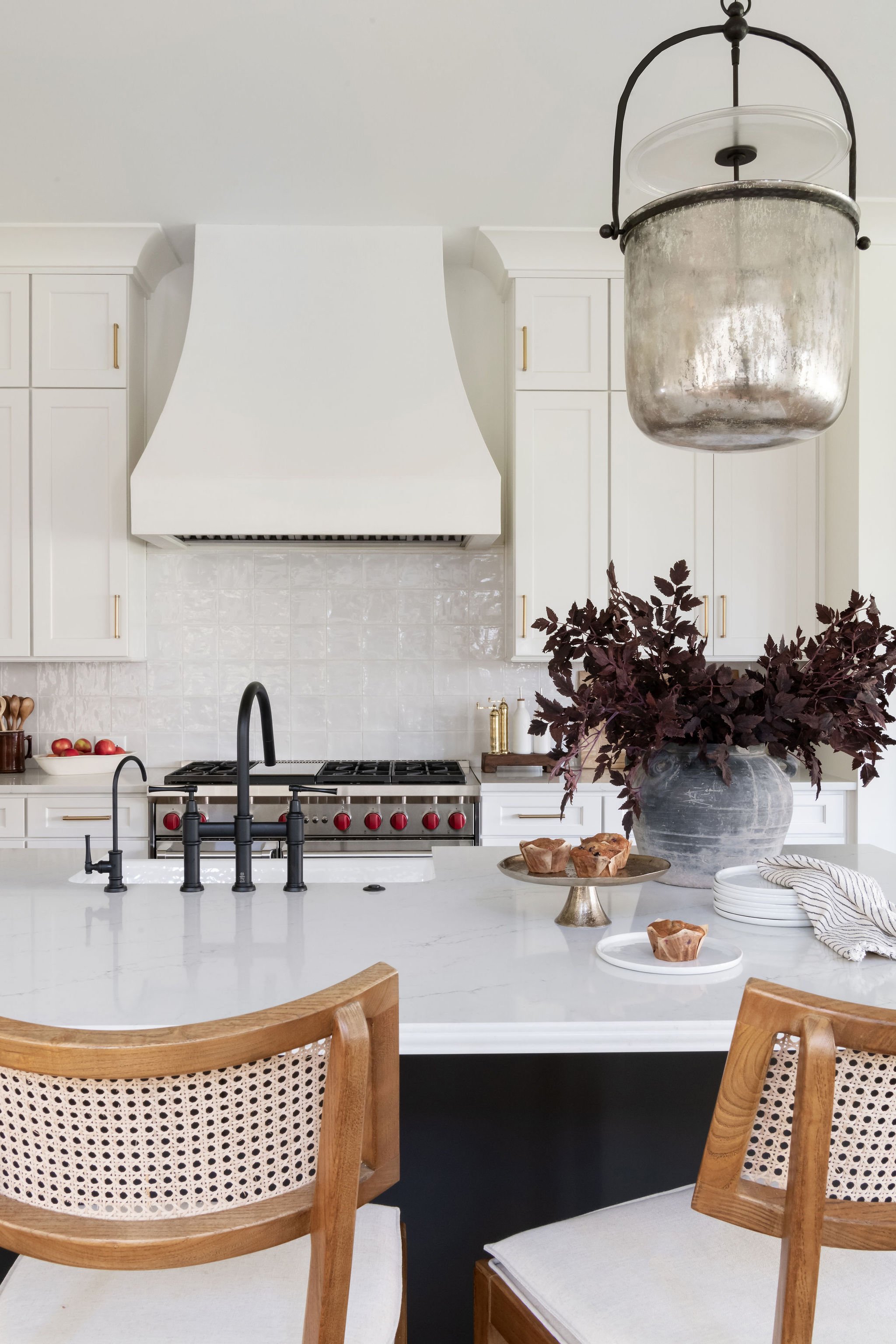 Modern kitchen with white cabinetry, black fixtures, a central island with a white marble countertop, a large hanging metallic pot, a dark leafy plant in a gray vase, and baked goods on plates.
