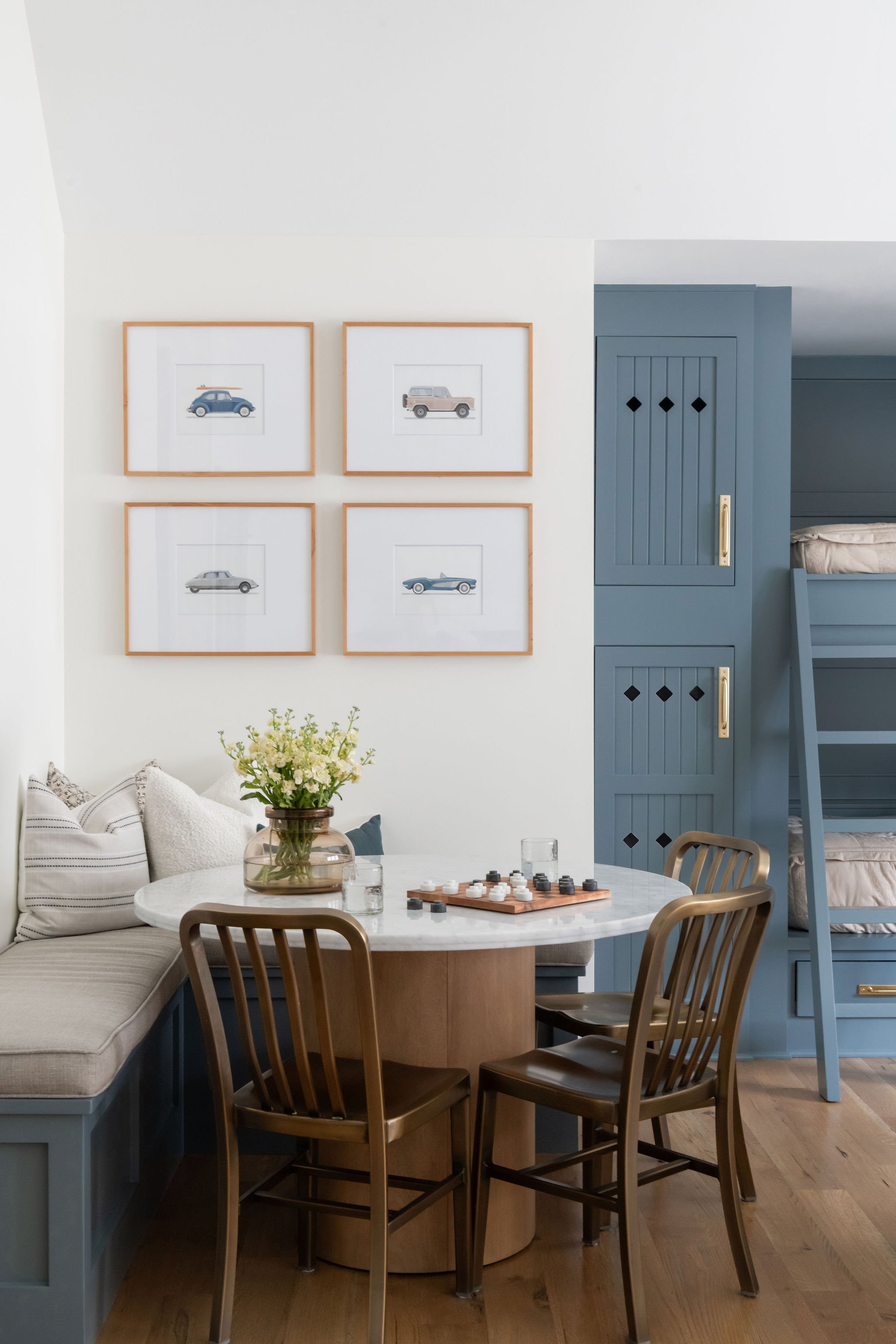 A cozy dining area with a round marble table, four wooden chairs, and a built-in corner bench with striped and textured pillows. On the table are glassware and a chess game. A vase of flowers sits on the corner of the table. The background features f