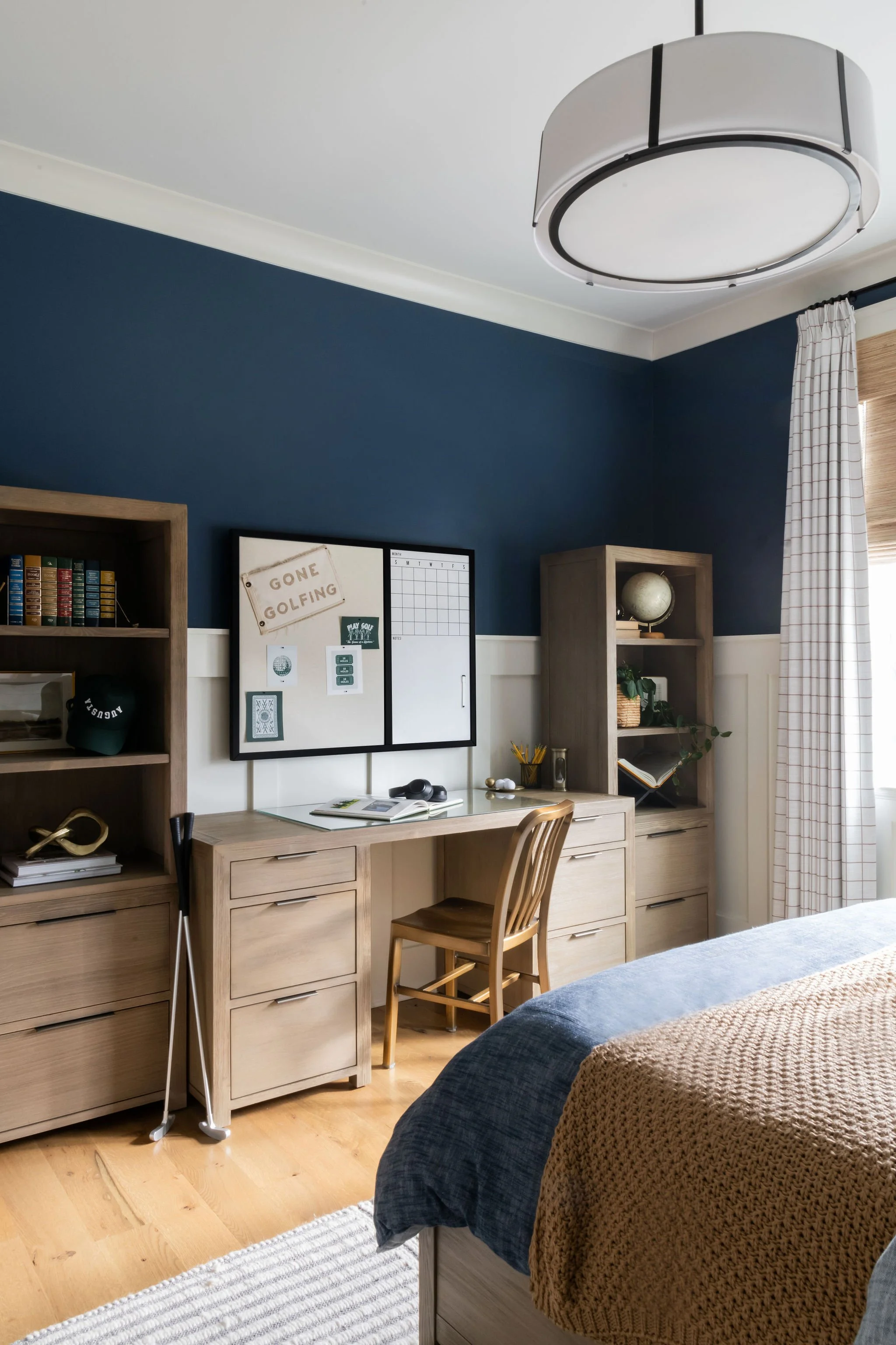 A bedroom corner with a wooden desk, a chair, and bookshelves against a dark blue wall. A whiteboard and corkboard are on the wall above the desk. There is a globe, plant, and decor items on the shelves. A window with curtains is on the right side, a