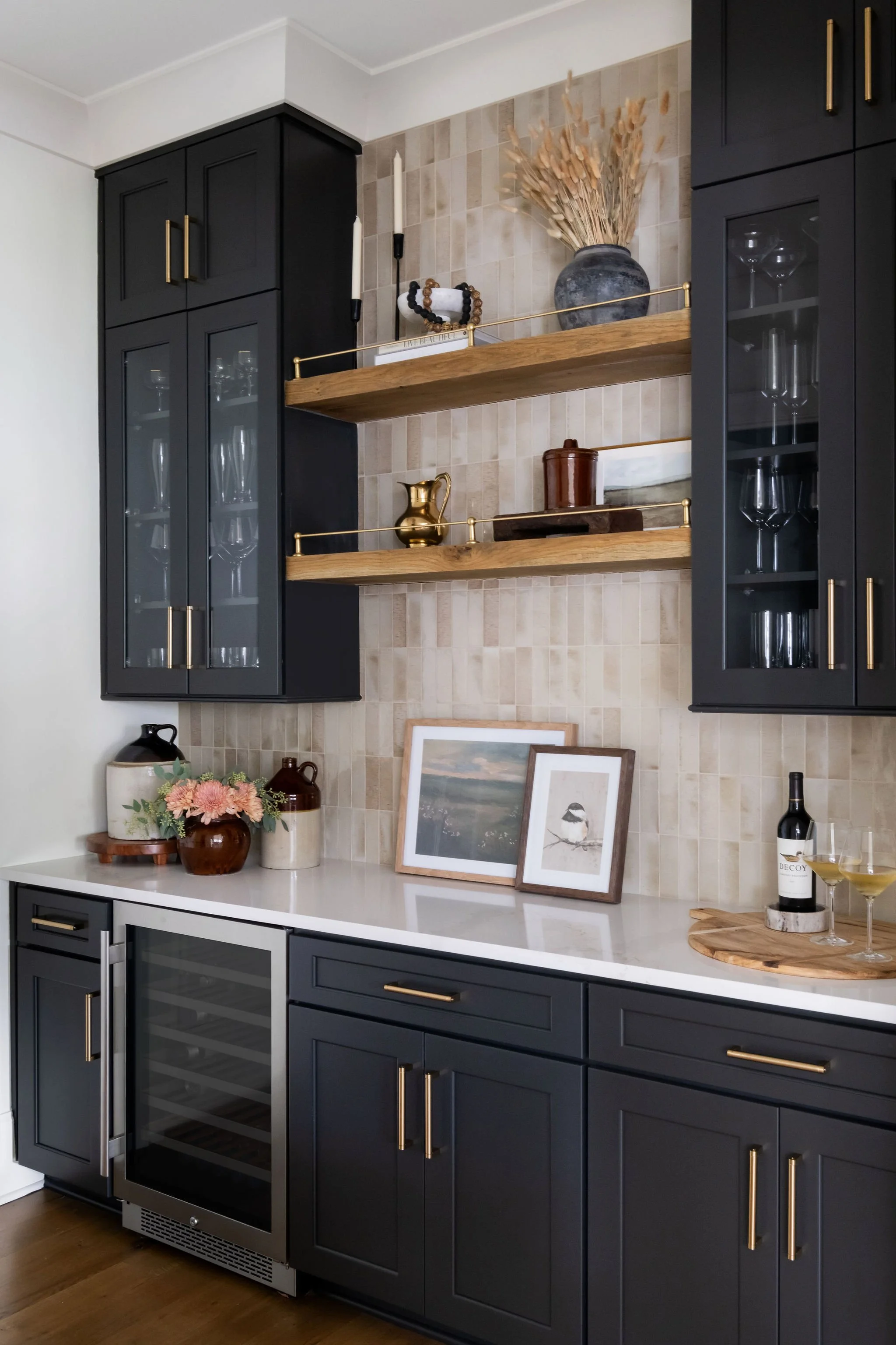 Decorative kitchen cabinet area with black cabinets, gold handles, and open wooden shelves holding vases, candles, and glassware; countertop displaying framed pictures, a flower arrangement, and a wine bottle with glasses.