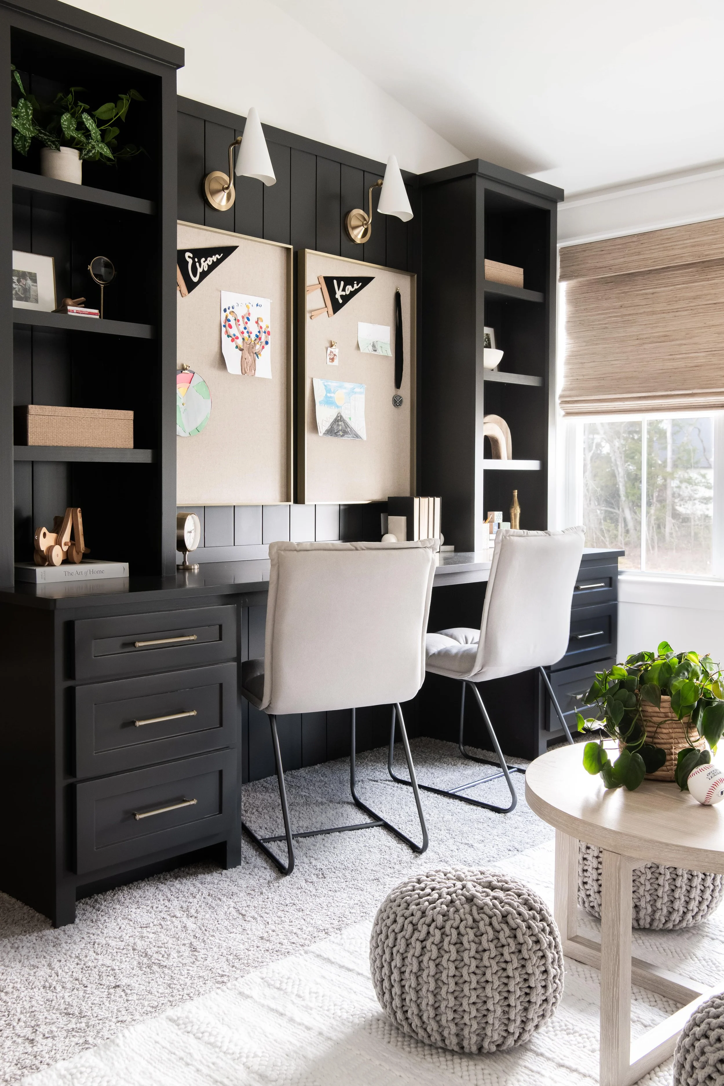Modern home office with black built-in desk, white chairs, bulletin board with children's artwork, beige window shades, a potted plant, and cozy decor.