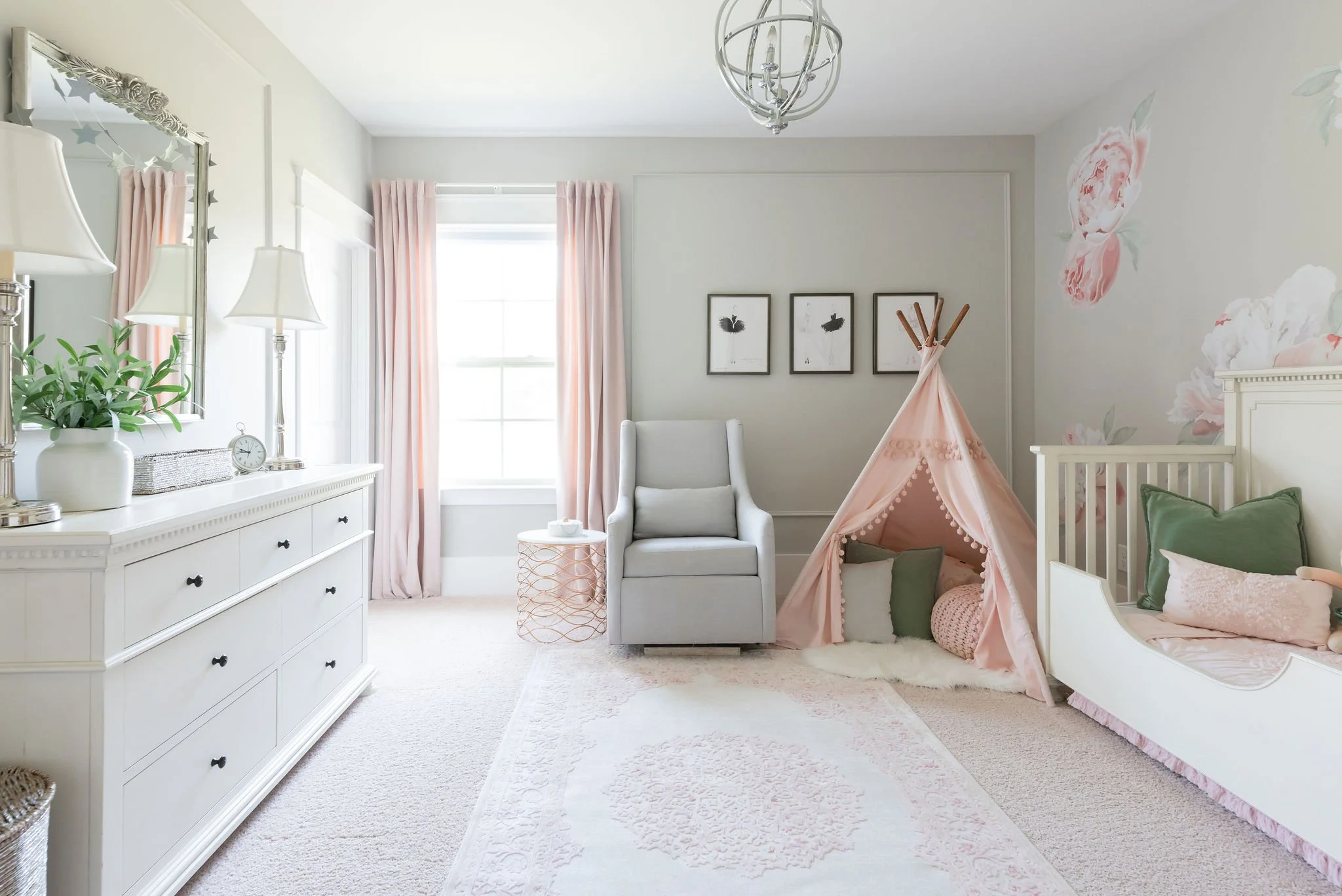 Children's bedroom with a white dresser, framed mirror, lamps, potted plant, gray armchair, pink tent, framed artwork, bed with green pillows, and floral wall decals.