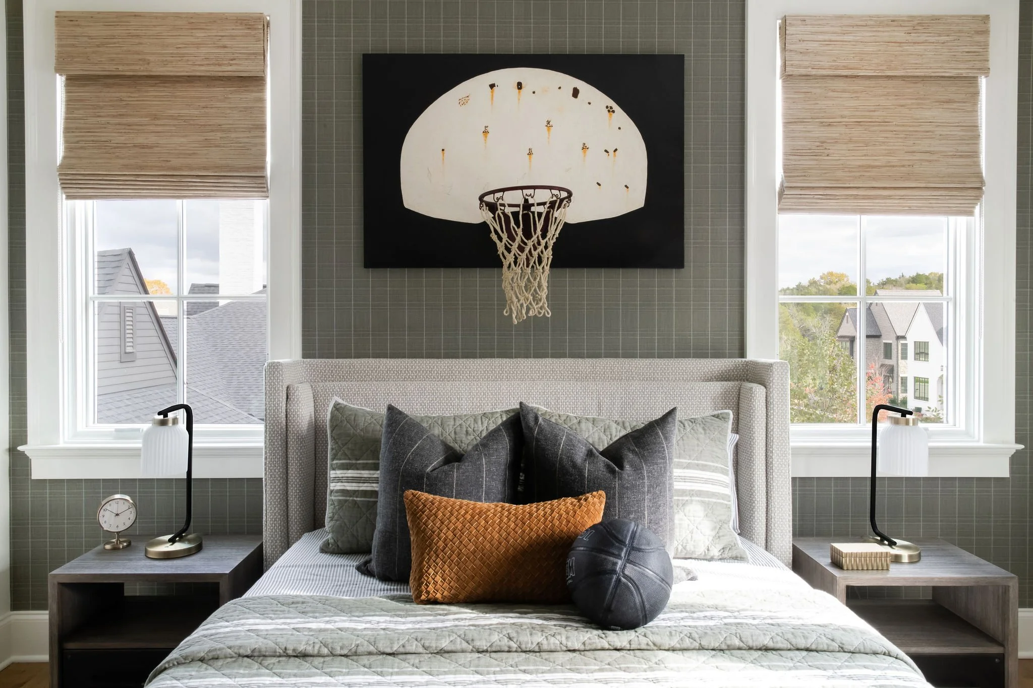 A bedroom with two large windows, a bed with decorative pillows, modern lamps on bedside tables, an abstract basketball hoop wall art, and a partial view of neighboring houses outside.
