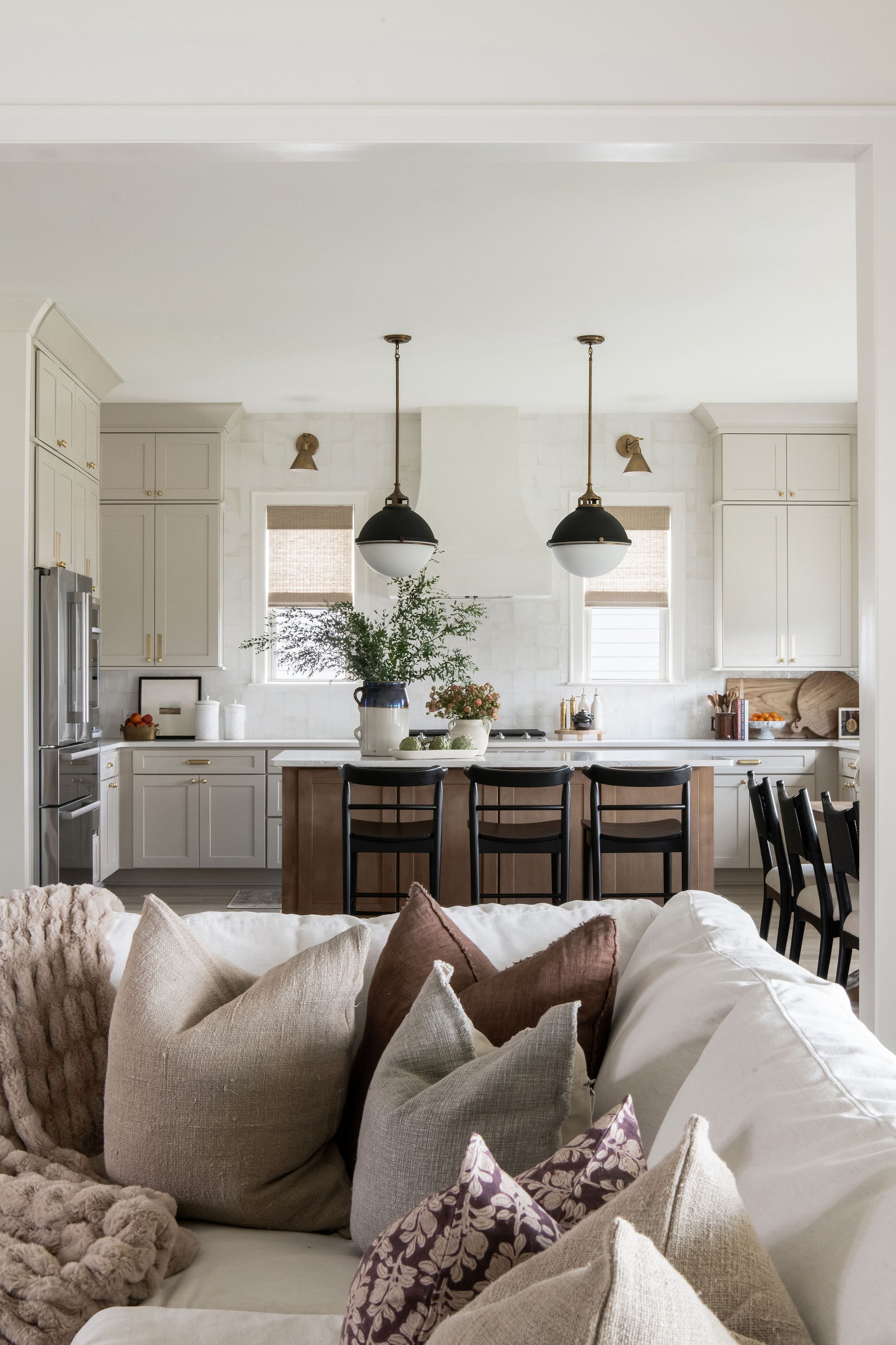 Living room with a view into a white kitchen, featuring black pendant lights, a kitchen island with black chairs, and beige pillows on a white sofa.