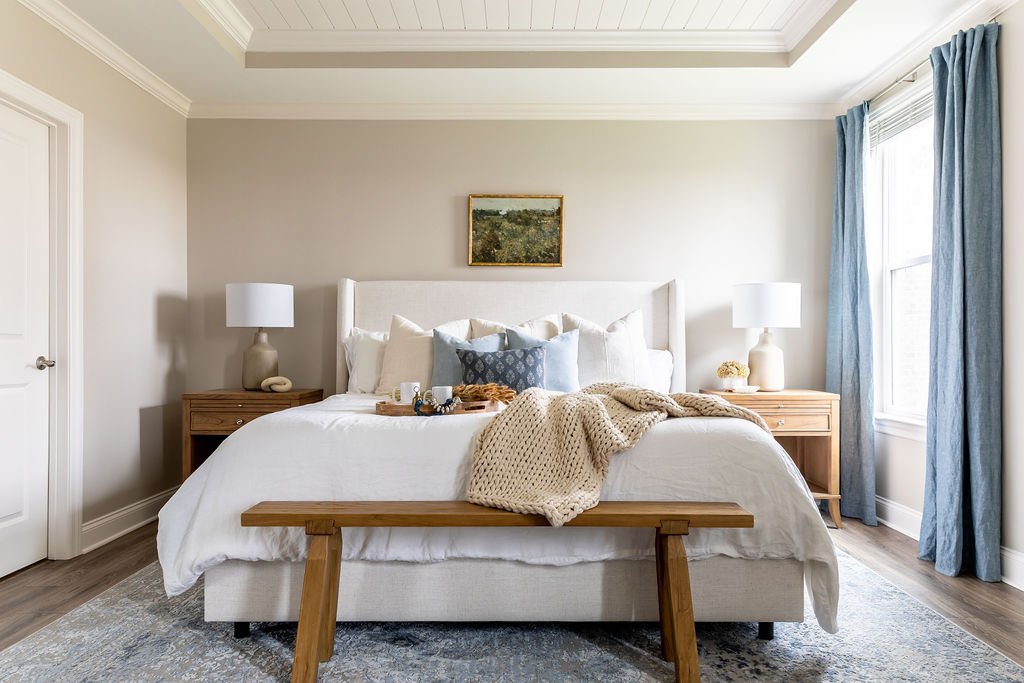 A neatly made bed in a bright bedroom with two wooden nightstands, white lamps, a small painting above the headboard, and light blue curtains.