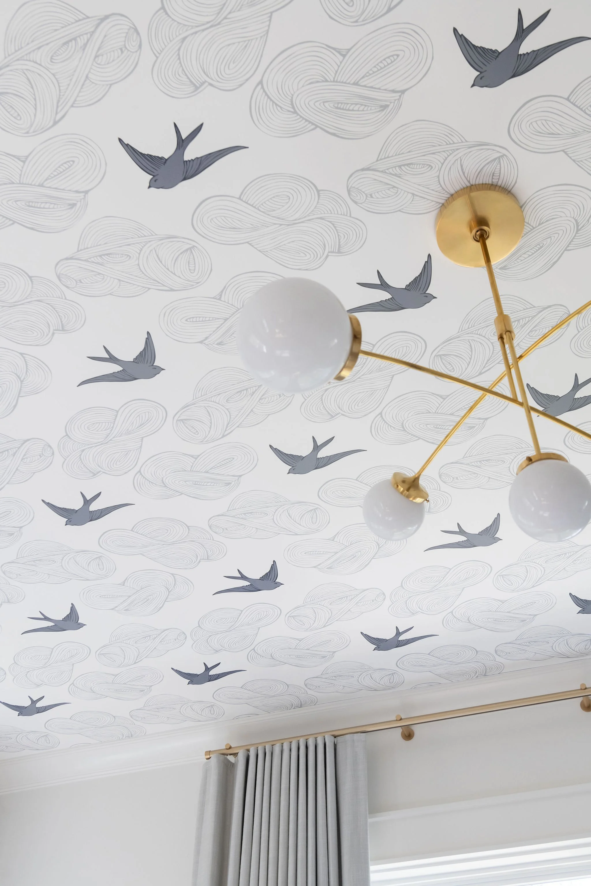 Ceiling with bird and cloud patterned wallpaper, modern gold chandelier with globe lights, and grey curtains.